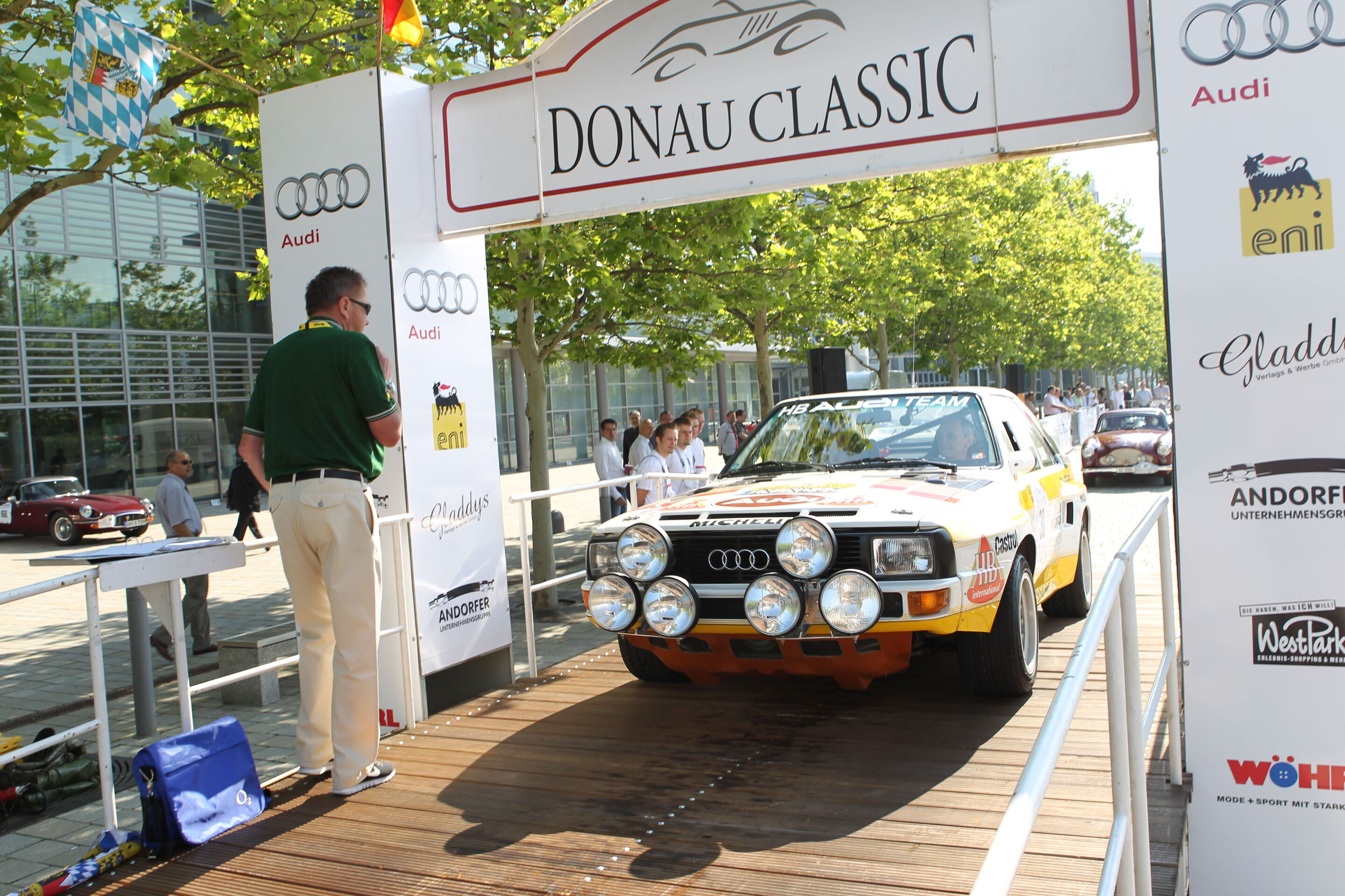 Donau Classic: Zieleinfahrt der Oldtimer-Rallye im Audi Forum Ingolstadt - Ein Audi fährt bei den Donau Classic durch die Zieleinfahrt am Audi Forum Ingolstadt. Zuschauer verfolgen das Ereignis am Streckenrand.