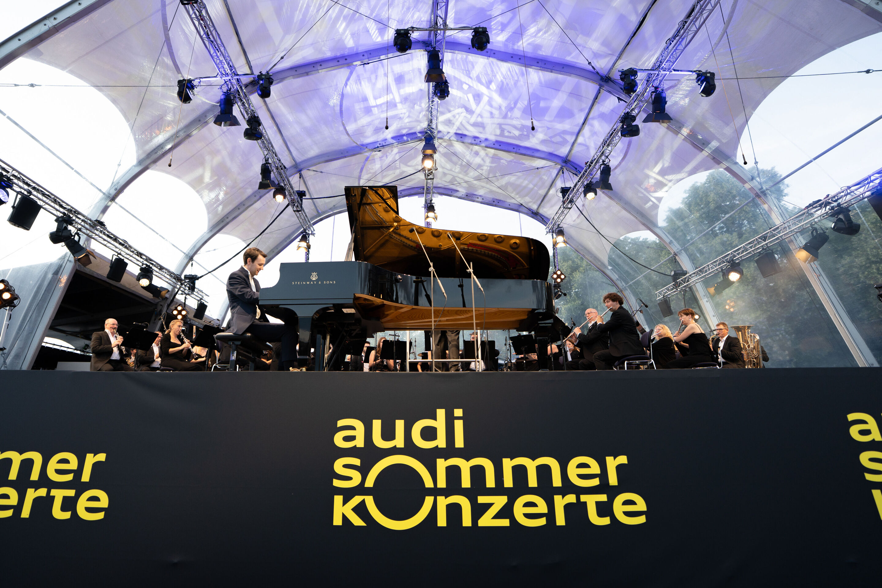 Klassik Open Air – Musical-Highlights mit der Audi Bläserphilharmonie - Beim Klassik Open Air im Klenzepark im Rahmen der Audi Sommerkonzerte spielt Florian Glemser auf der Bühne auf dem Piano; um ihn herum spielen Mitglieder der Audi Bläserphilharmonie...