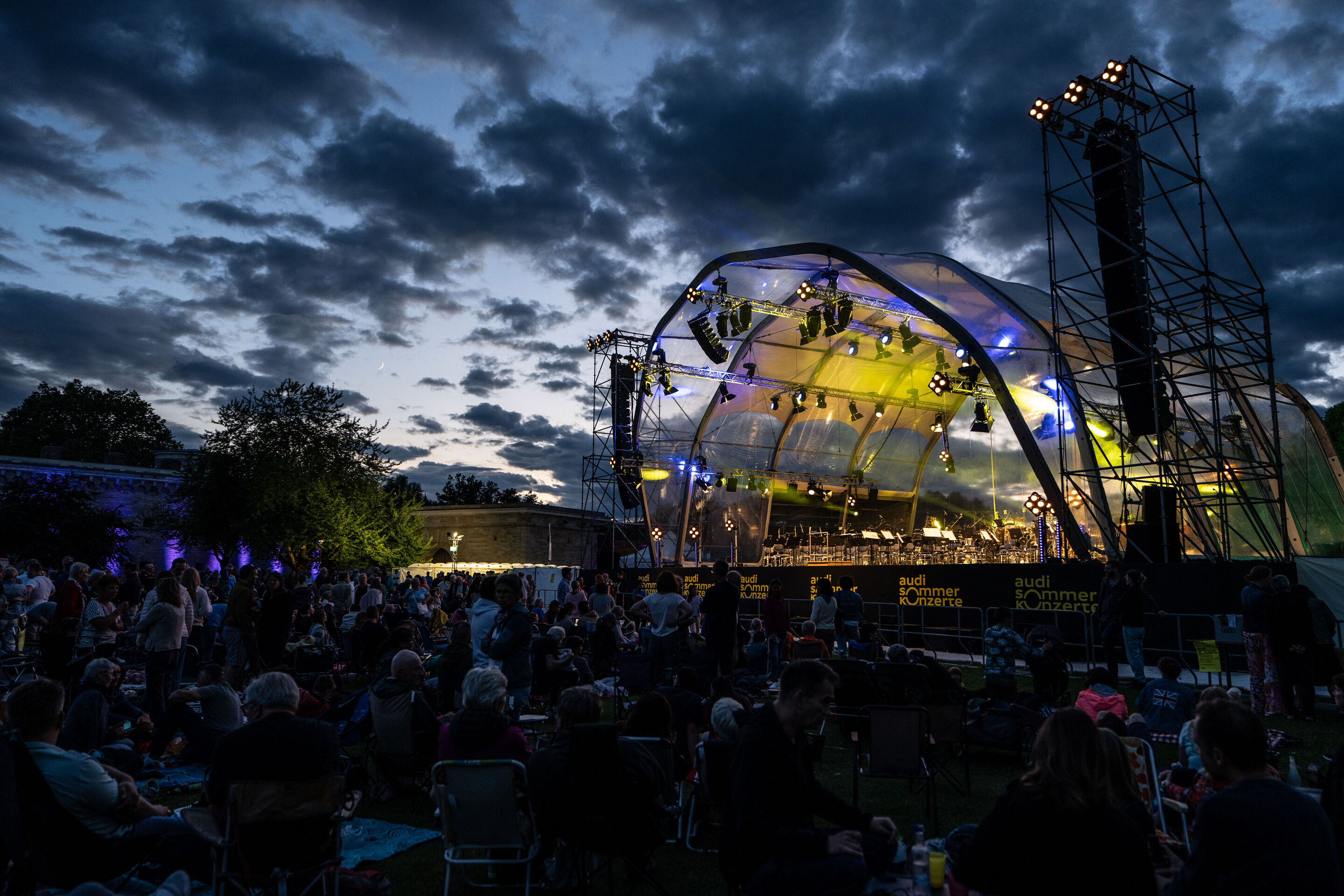 Klassik Open Air – Musical-Highlights mit der Audi Bläserphilharmonie - Beim Klassik Open Air im Klenzepark im Rahmen der Audi Sommerkonzerte sitzt das Publikum in der Abenddämmerung vor beleuchteten Bühne.