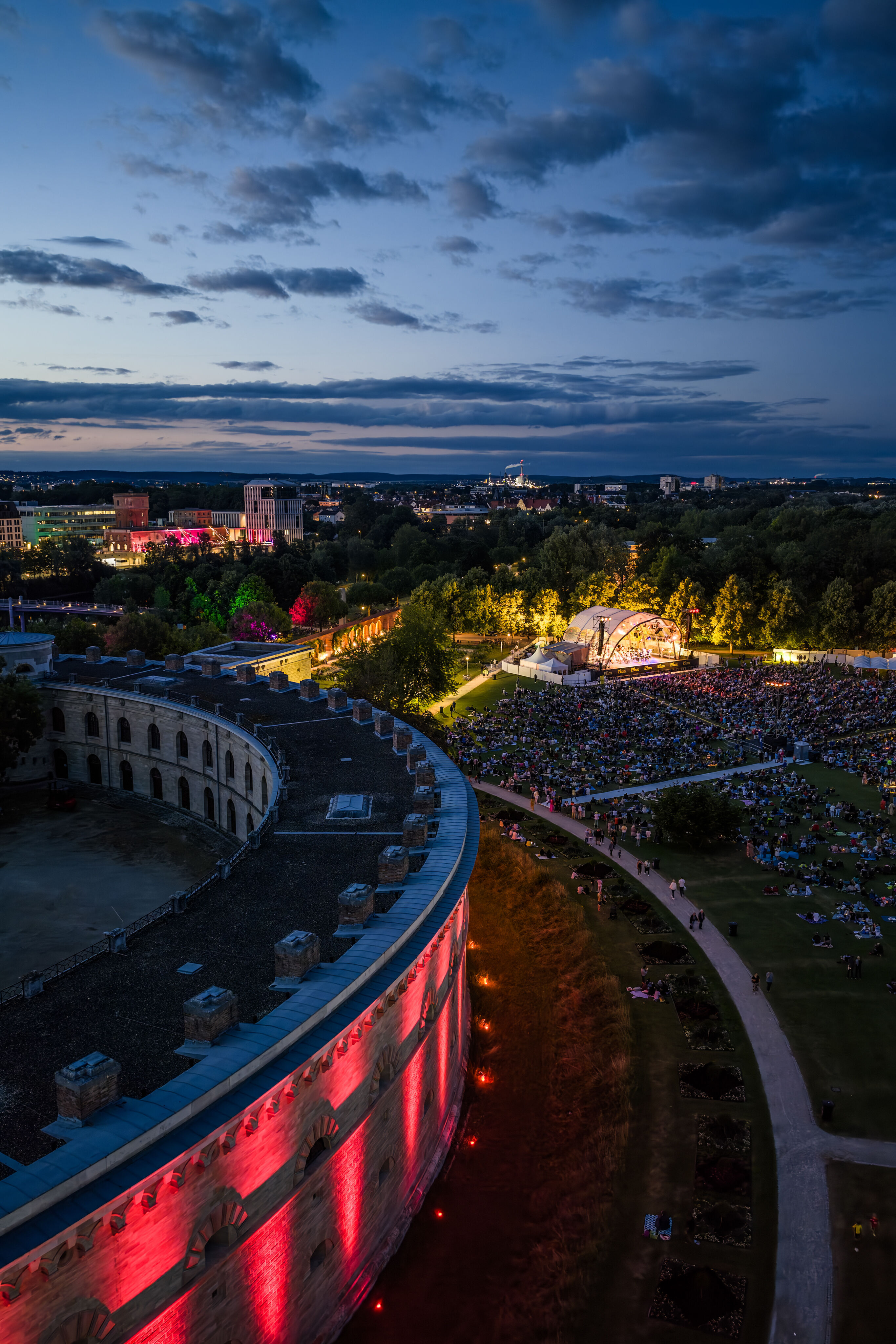Klassik Open Air – Musical-Highlights mit der Audi Bläserphilharmonie - Blick aus der Vogelperspektive in der Abenddämmerung auf das Klassik Open Air mit der Audi Bläserphilharmonie im Rahmen der Audi Sommerkonzerte im Klenzepark in Ingolstadt; im Vordergrund...