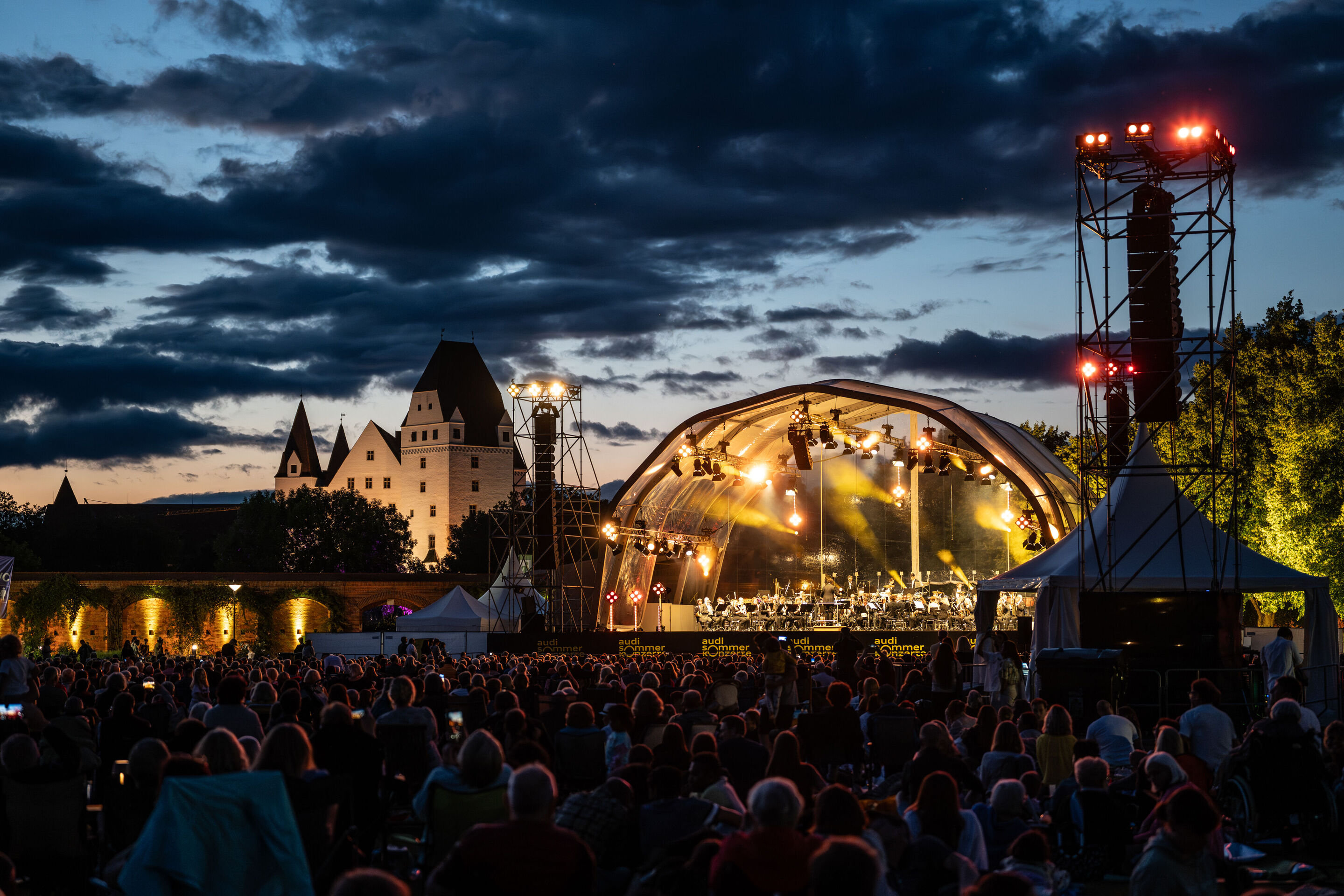 Klassik Open Air – Musical-Highlights mit der Audi Bläserphilharmonie - Beim Klassik Open Air im Klenzepark im Rahmen der Audi Sommerkonzerte sitzt das Publikum in der Abenddämmerung vor der beleuchteten Bühne.