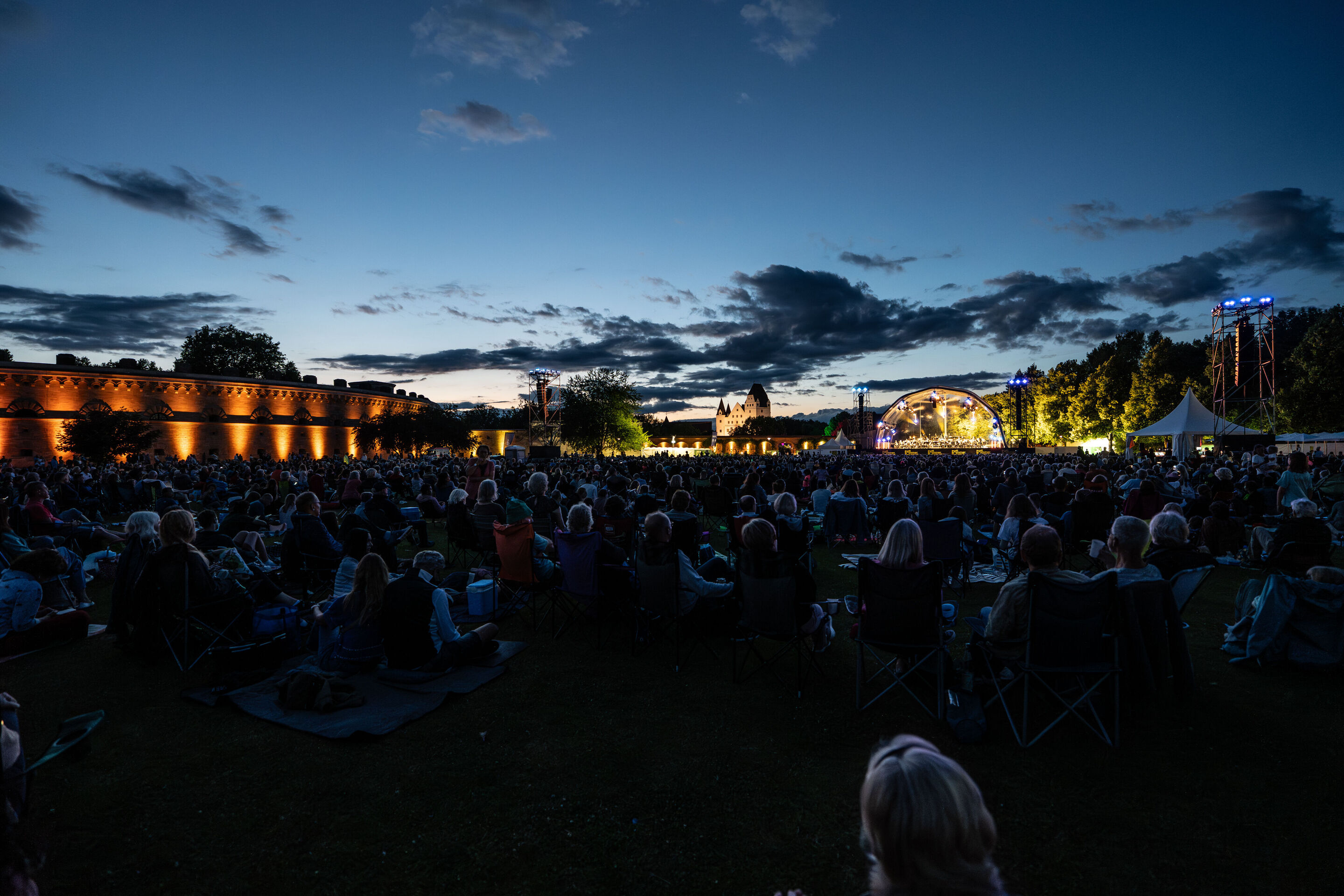 Klassik Open Air – Musical-Highlights mit der Audi Bläserphilharmonie - Beim Klassik Open Air im Klenzepark im Rahmen der Audi Sommerkonzerte sitzt das Publikum in der Abenddämmerung vor der beleuchteten Bühne, auf der linken Seite ist das beleuchtete...