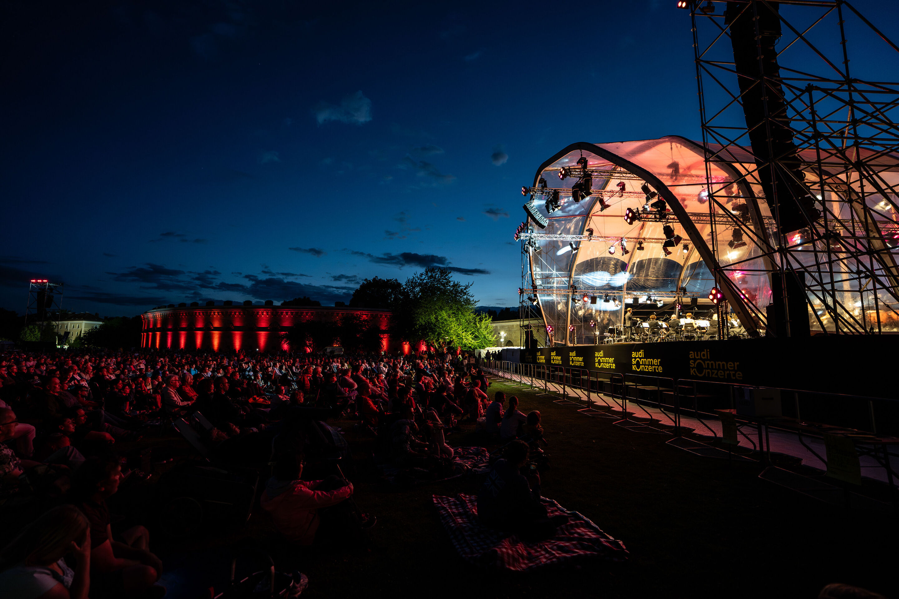 Klassik Open Air – Musical-Highlights mit der Audi Bläserphilharmonie - Beim Klassik Open Air im Klenzepark im Rahmen der Audi Sommerkonzerte sitzt das Publikum in der Abenddämmerung vor der beleuchteten Bühne, auf der linken Seite ist das beleuchtete...