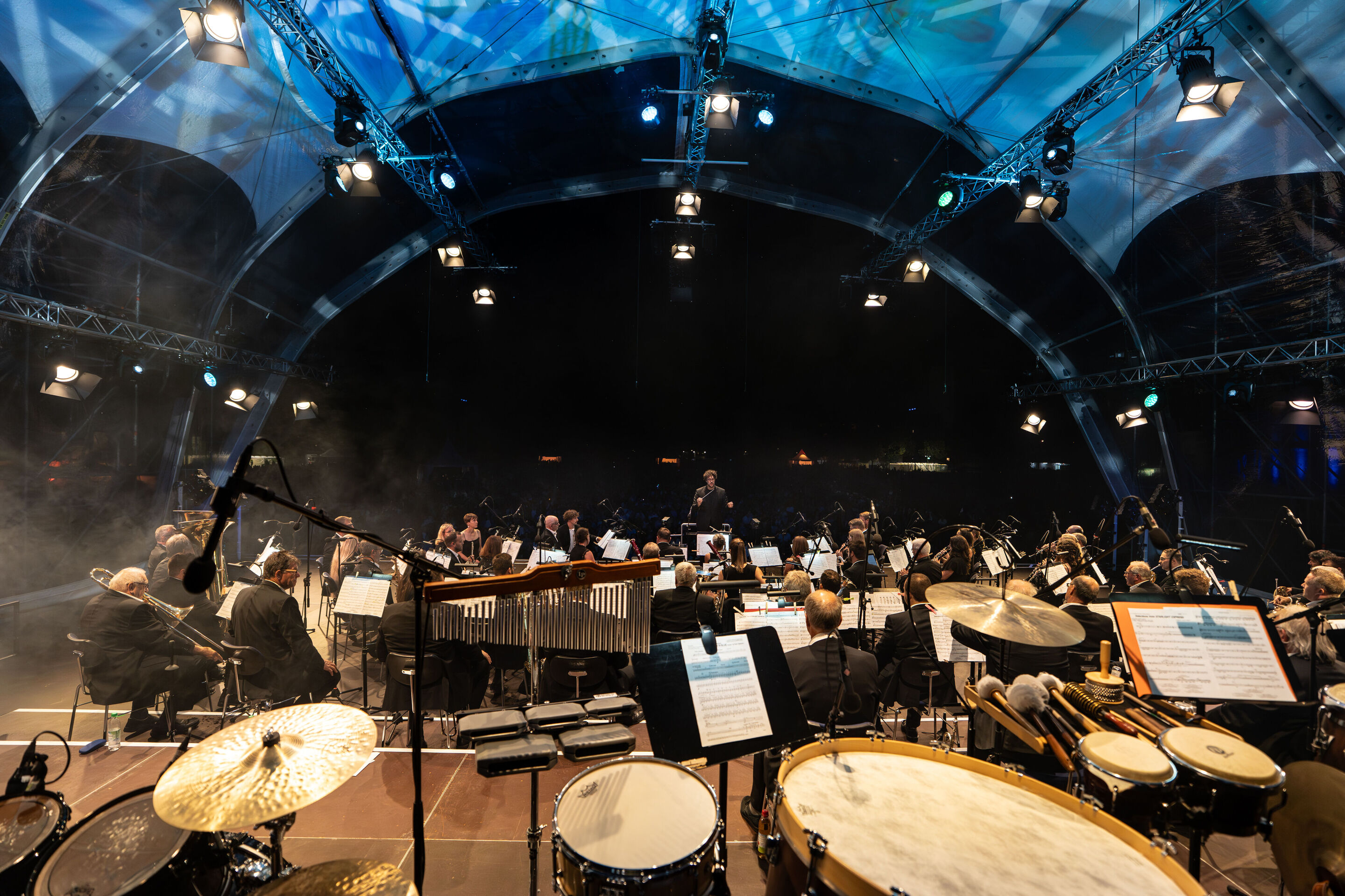 Klassik Open Air – Musical-Highlights mit der Audi Bläserphilharmonie - Blick von der Bühne in Richtung Publikum: Pietro Sarno dirigiert beim Klassik Open Air im Klenzepark im Rahmen der Audi Sommerkonzerte die Audi Bläserphilharmonie.