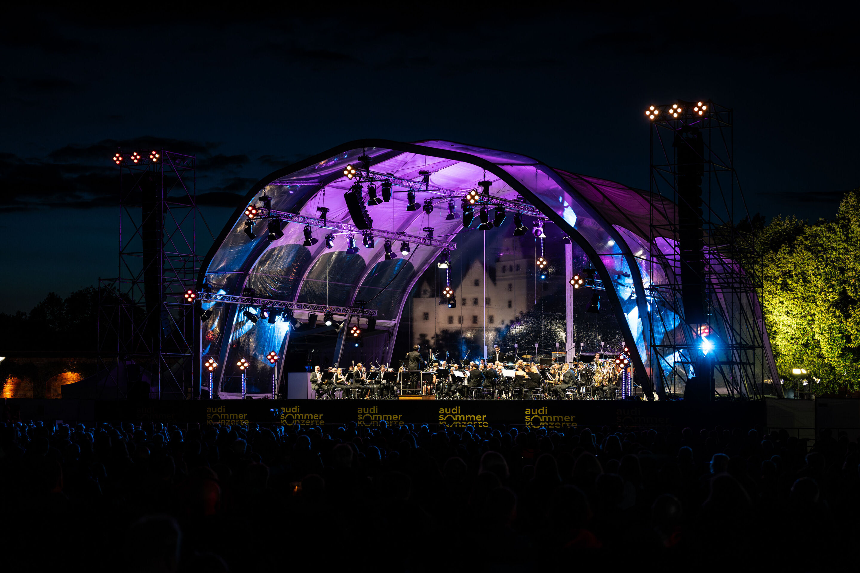 Klassik Open Air – Musical-Highlights mit der Audi Bläserphilharmonie - Beim Klassik Open Air im Klenzepark im Rahmen der Audi Sommerkonzerte sitzt das Publikum abends vor der beleuchteten Bühne.