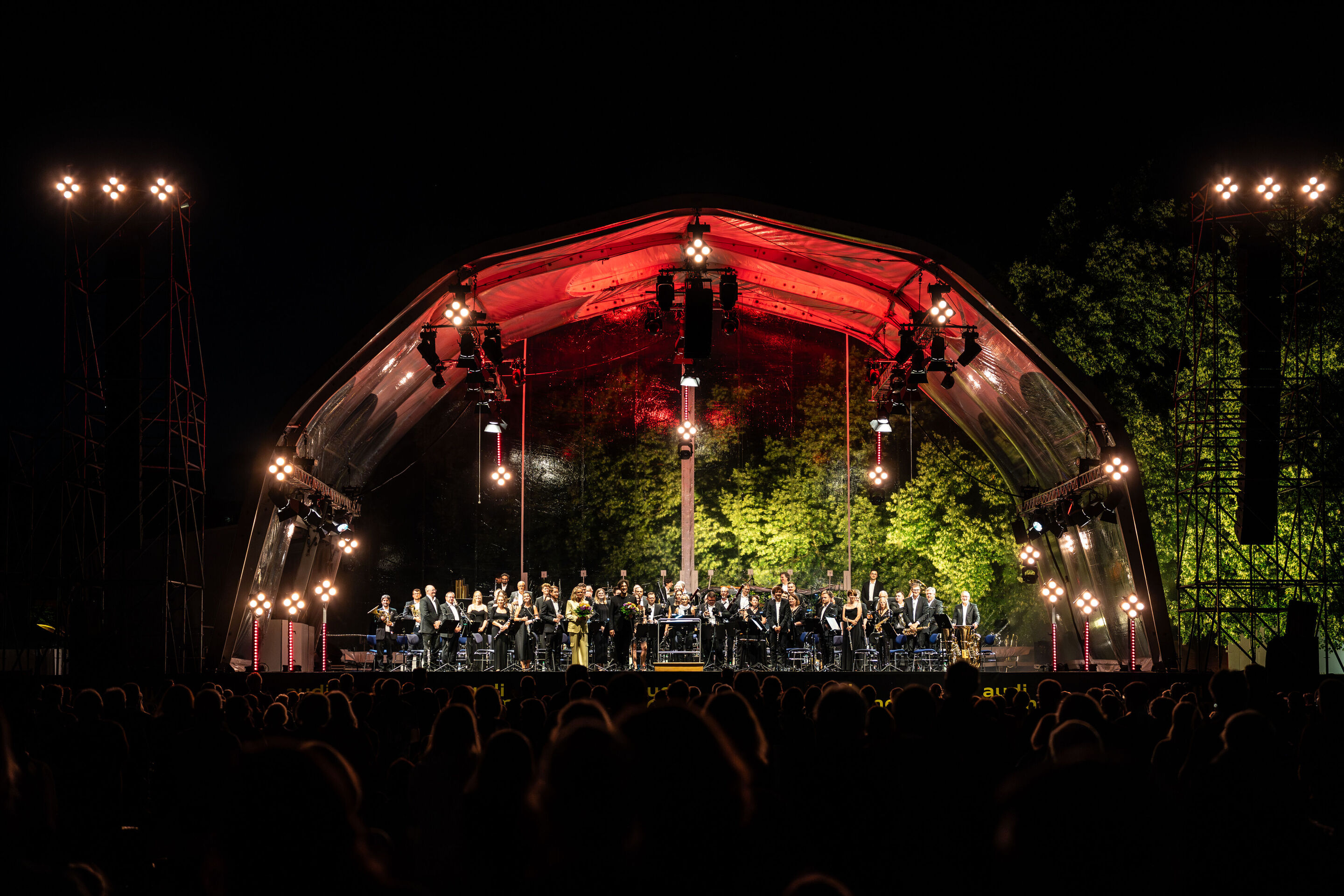 Klassik Open Air – Musical-Highlights mit der Audi Bläserphilharmonie - Die Audi Bläserphilharmonie, Dirigent Pietro Sarno und Moderatorin Annekatrin Hentschel stehen beim Klassik Open Air im Klenzepark im Rahmen der Audi Sommerkonzerte auf der beleuchteten...