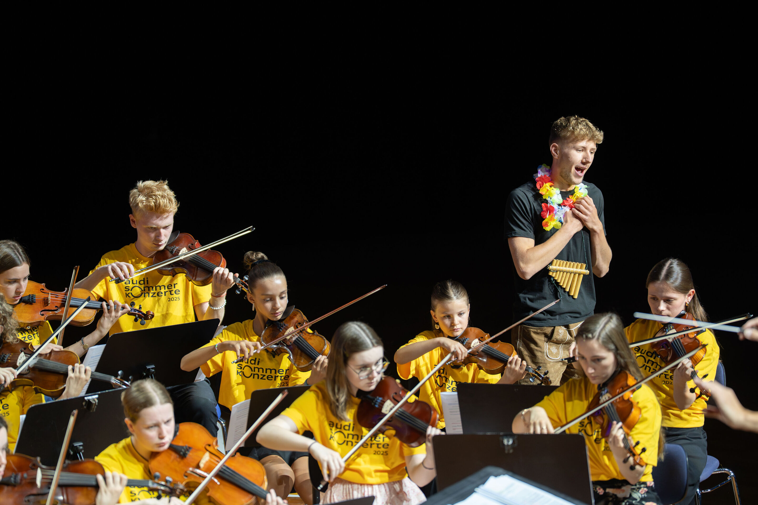 Öffentlicher Workshop mit Schülerinnen und Schülern der Region und dem Aurora Orchestra - Schüler Lukas Scheider (Q12, Gymnasium Wolnzach) singt rechts auf der Bühne des Festsaals Ingolstadt. Vor ihm sind musizierende Schülerinnen und Schüler zu sehen.
