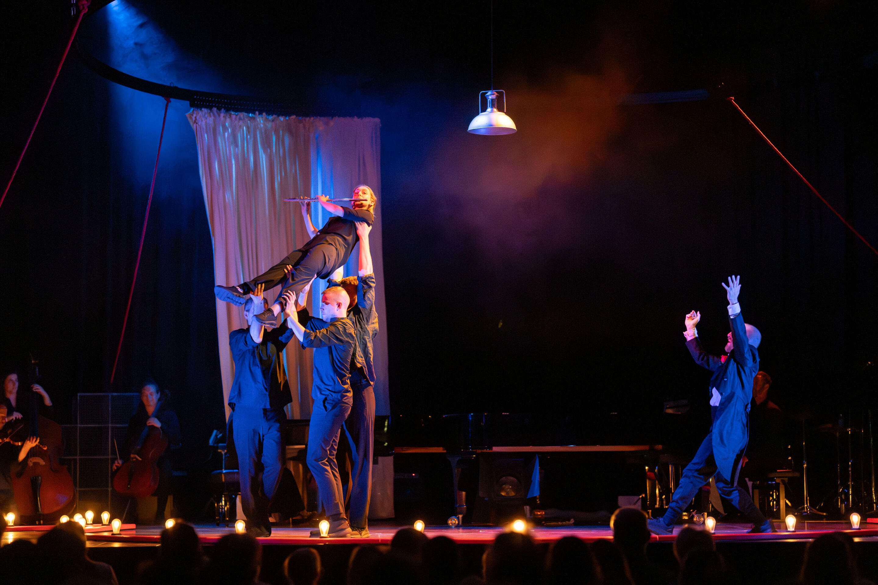 “Carnival”: Theatrical concert for families about the fabulous world of instruments - On the stage of the Festival Hall Ingolstadt, three members of the Aurora Orchestra lift a flutist into the air. To the right is dancer Christopher Akrill.