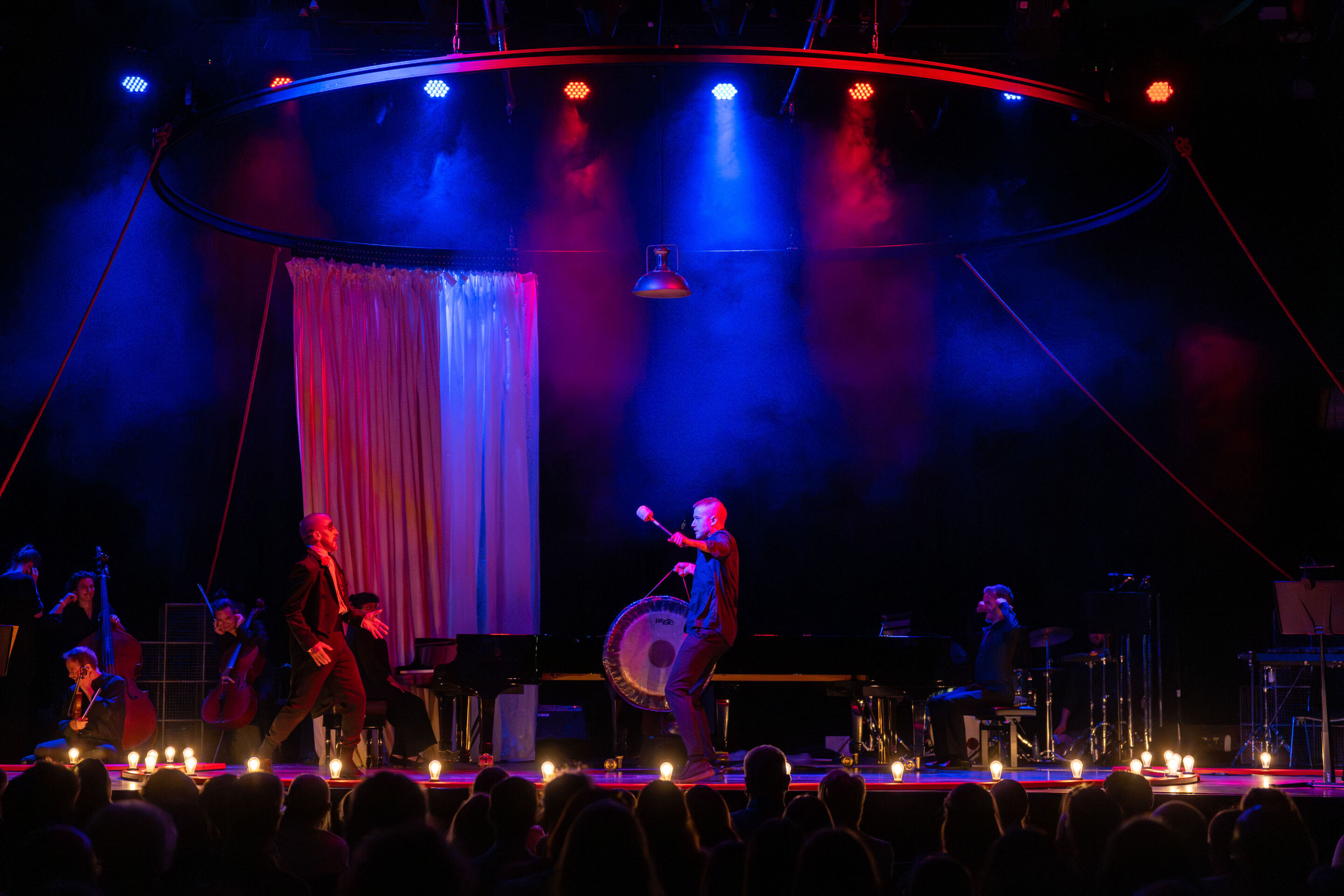 “Carnival”: Theatrical concert for families about the fabulous world of instruments - On the stage of the Festival Hall Ingolstadt, a member of the Aurora Orchestra stands holding a symphonic gong in front of dancer Christopher Akrill, who is positioned...