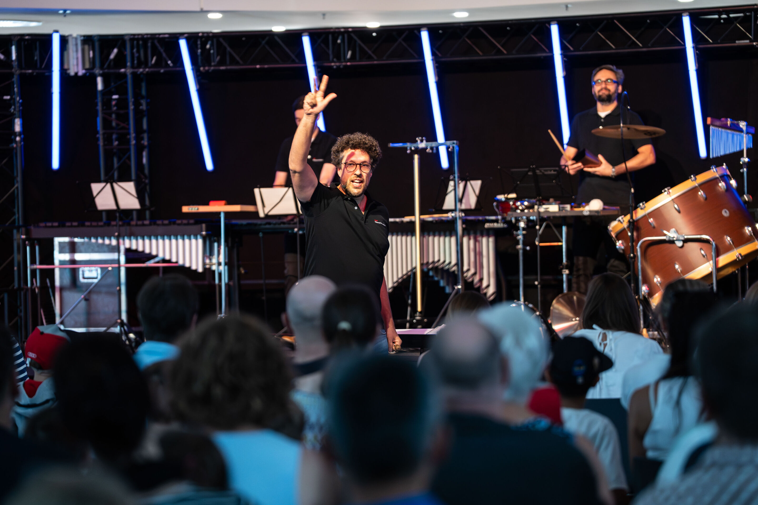 “ADUI und die Kraft der magischen Ringe”: children’s concert with the Audi Philharmonic Wind Orchestra - Conductor Pietro Sarno,on stage of the H6 building of the Ingolstadt site, looks at the audience and holds up three fingers.