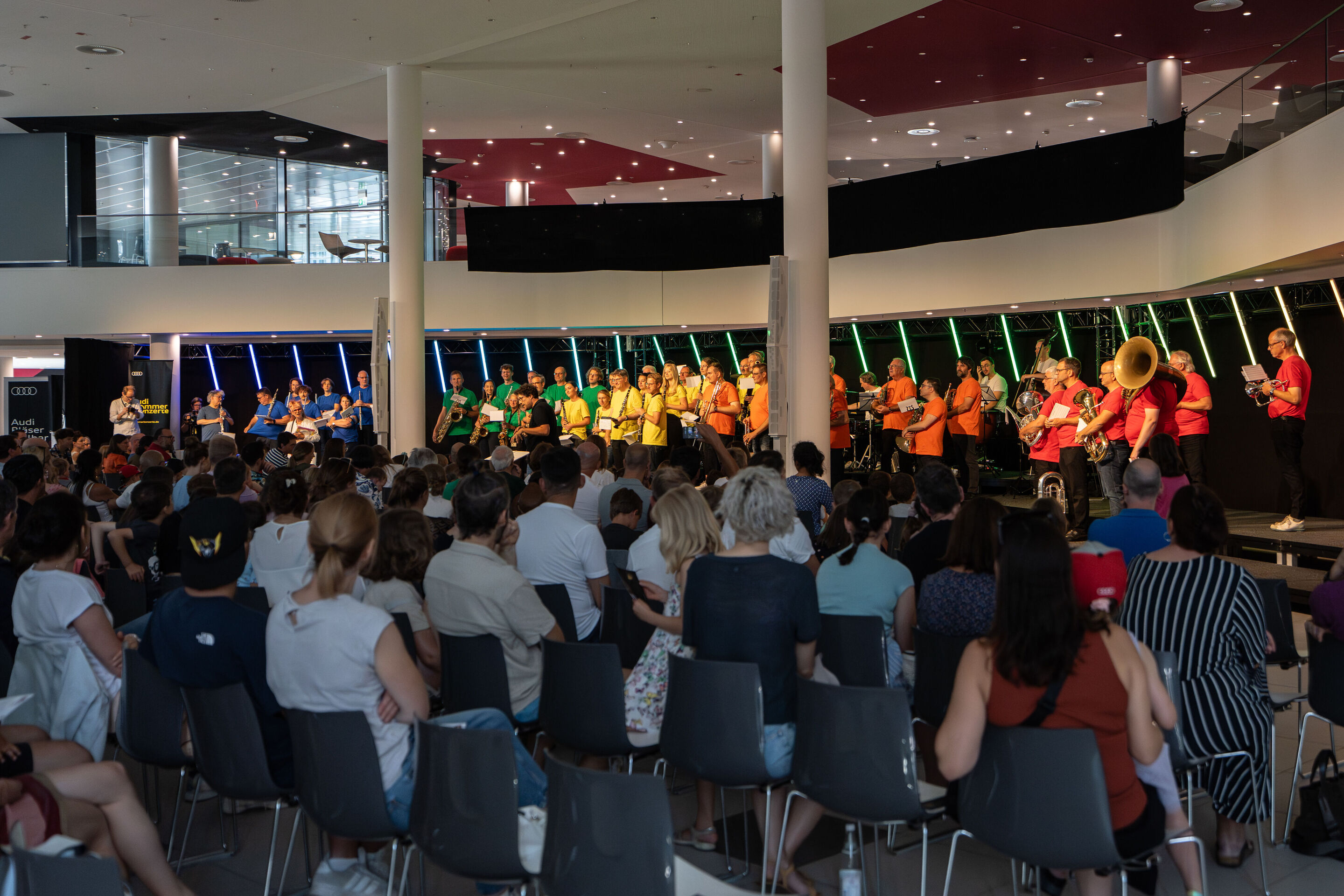 “ADUI und die Kraft der magischen Ringe”: children’s concert with the Audi Philharmonic Wind Orchestra - The musicians of the Audi Philharmonic Wind Orchestra are on the stage of the H6 building at the Ingolstadt site, wearing colorful T-shirts with conductor...