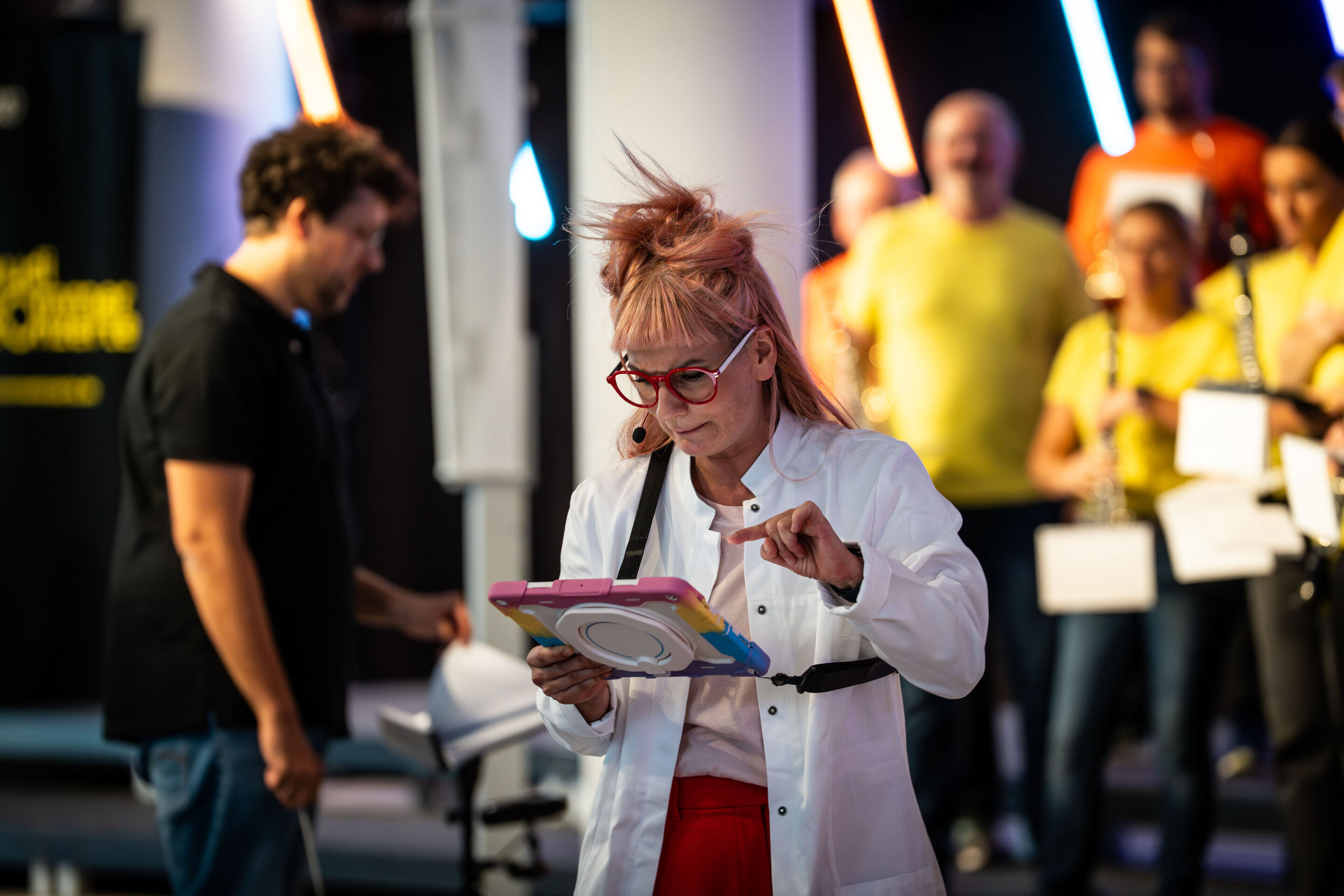 “ADUI und die Kraft der magischen Ringe”: children’s concert with the Audi Philharmonic Wind Orchestra - Olivia Wendt is seen as a researcher wearing a white lab coat, with disheveled hair and a tablet in her hands. In the blurred background are conductor...