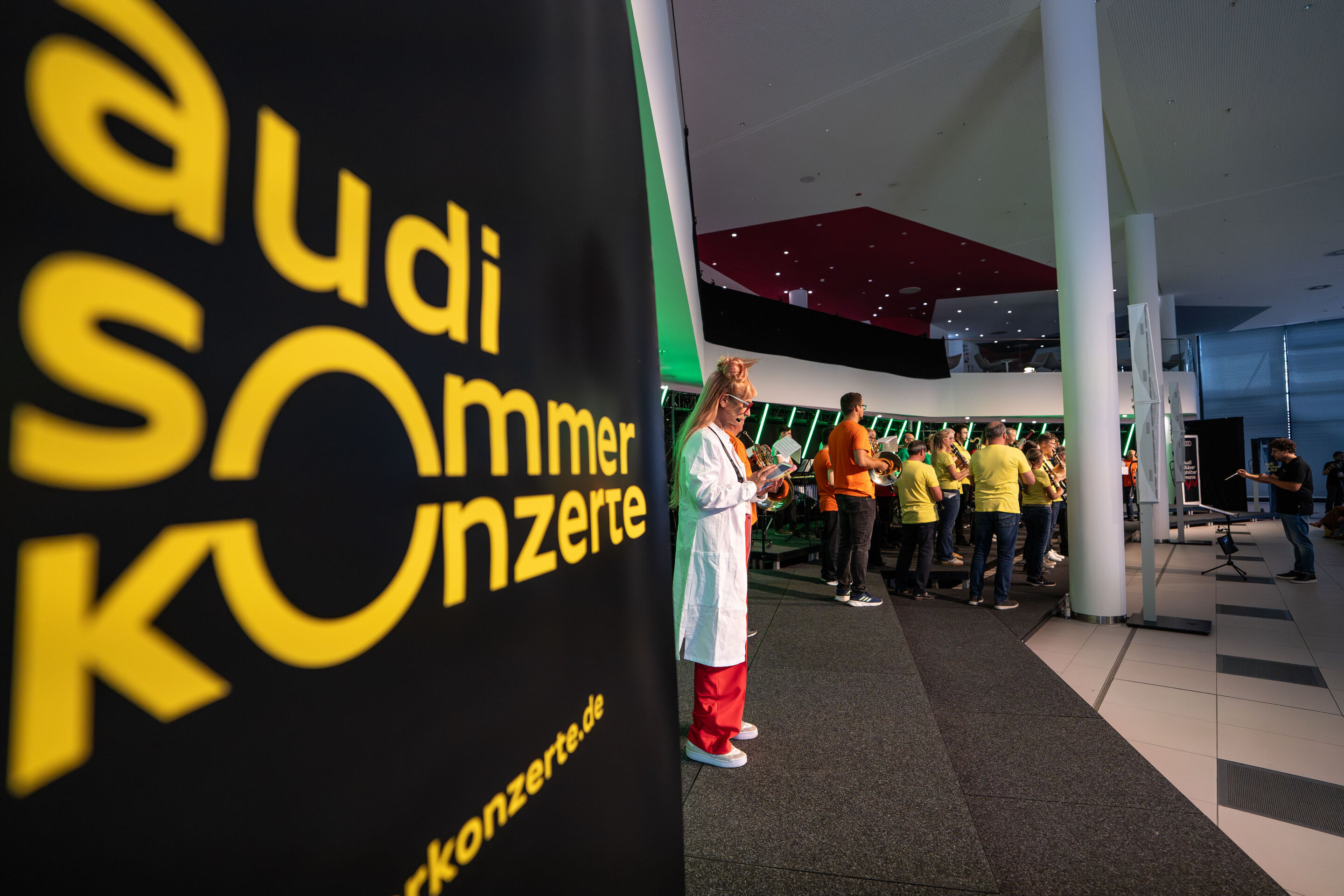“ADUI und die Kraft der magischen Ringe”: children’s concert with the Audi Philharmonic Wind Orchestra - Olivia Wendt, dressed as a researcher, stands in the foreground while the orchestra performs in the background. The Audi Summer Concerts logo is displayed...