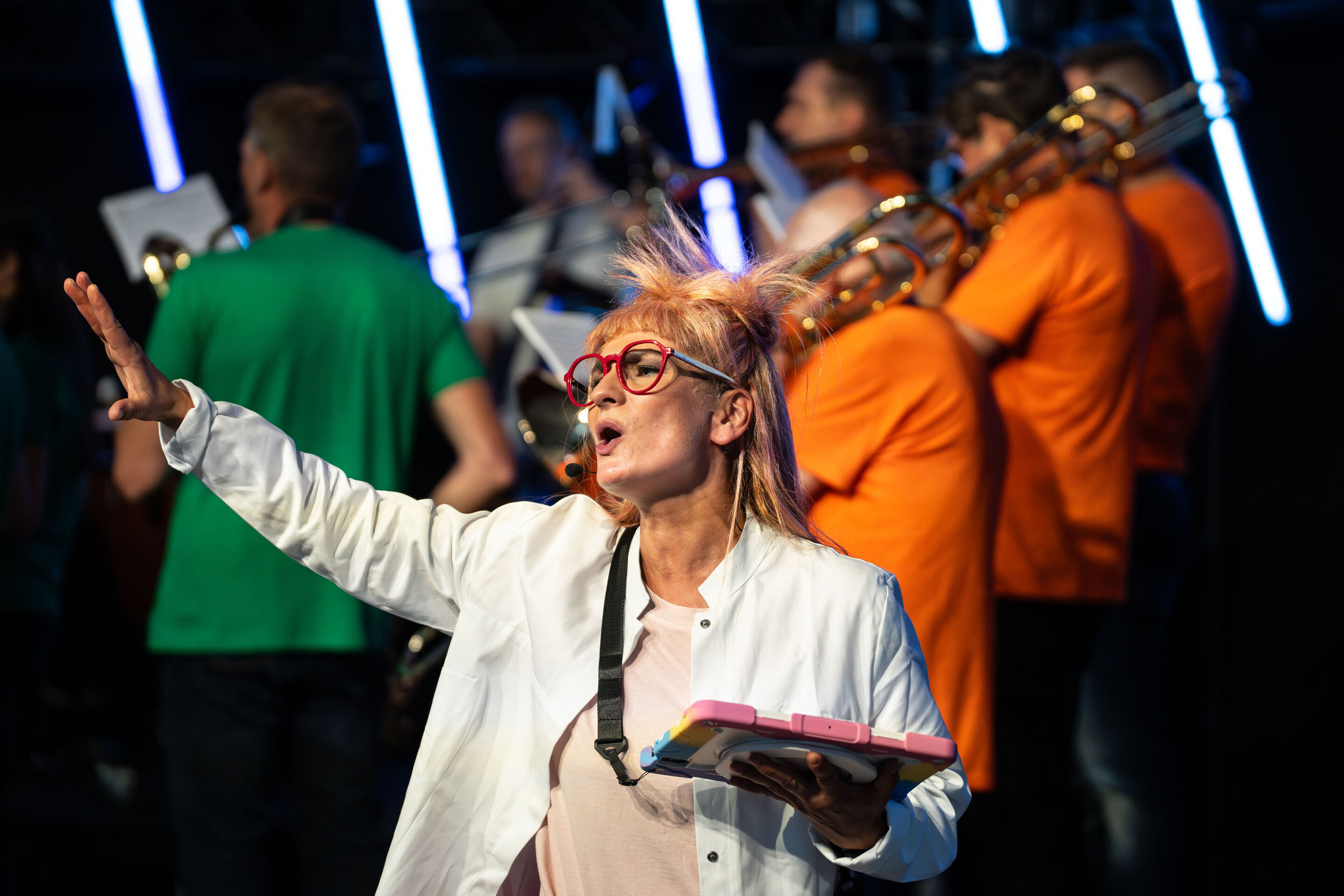 “ADUI und die Kraft der magischen Ringe”: children’s concert with the Audi Philharmonic Wind Orchestra - Olivia Wendt stands on stage dressed in a white lab coat as a researcher, holding a tablet in her hand while gesturing. In the background, the Audi...