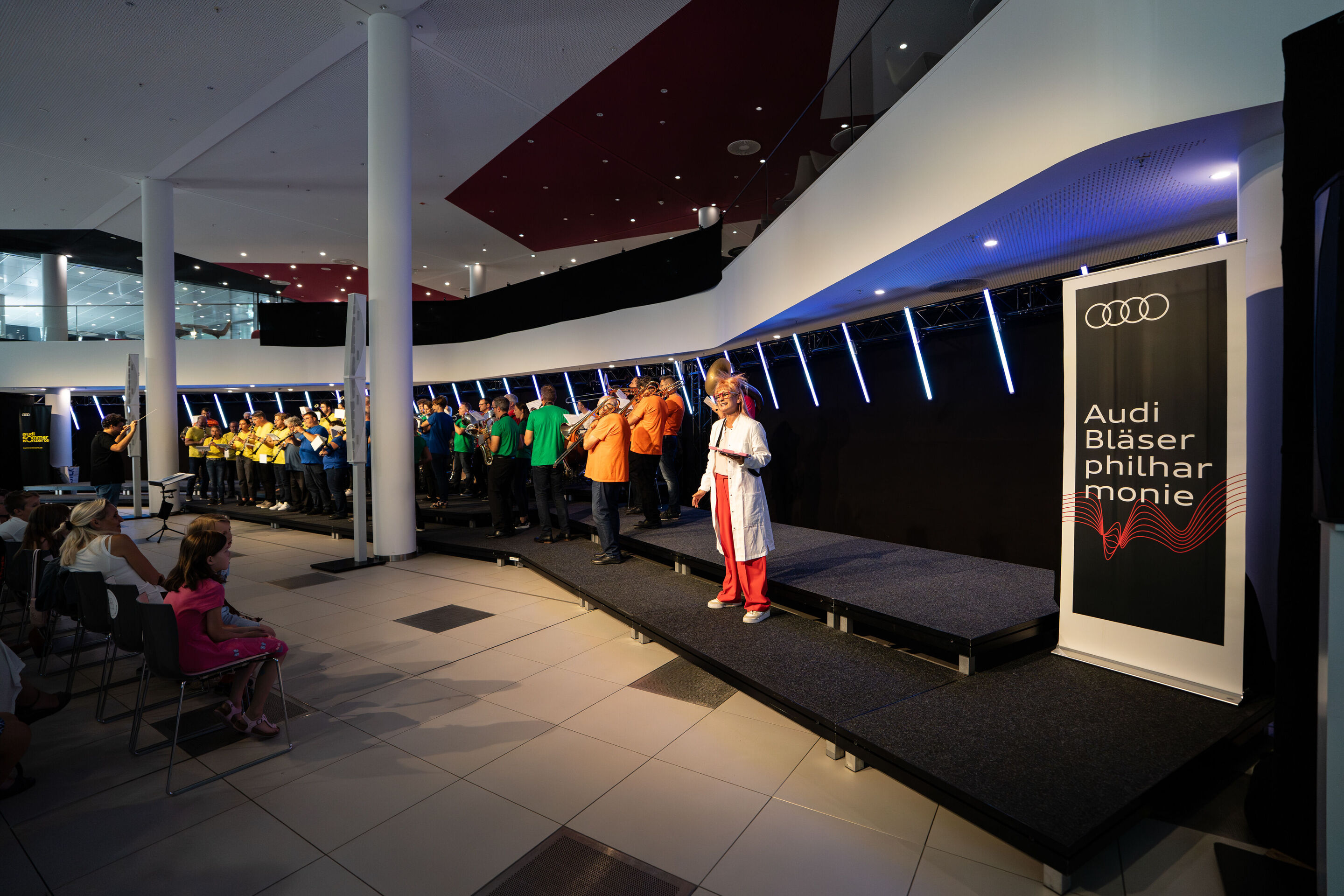 “ADUI und die Kraft der magischen Ringe”: children’s concert with the Audi Philharmonic Wind Orchestra - On stage stands Olivia Wendt dressed as a scientist. In the background, the Audi Philharmonic Wind Orchestra performs under the direction of conductor...
