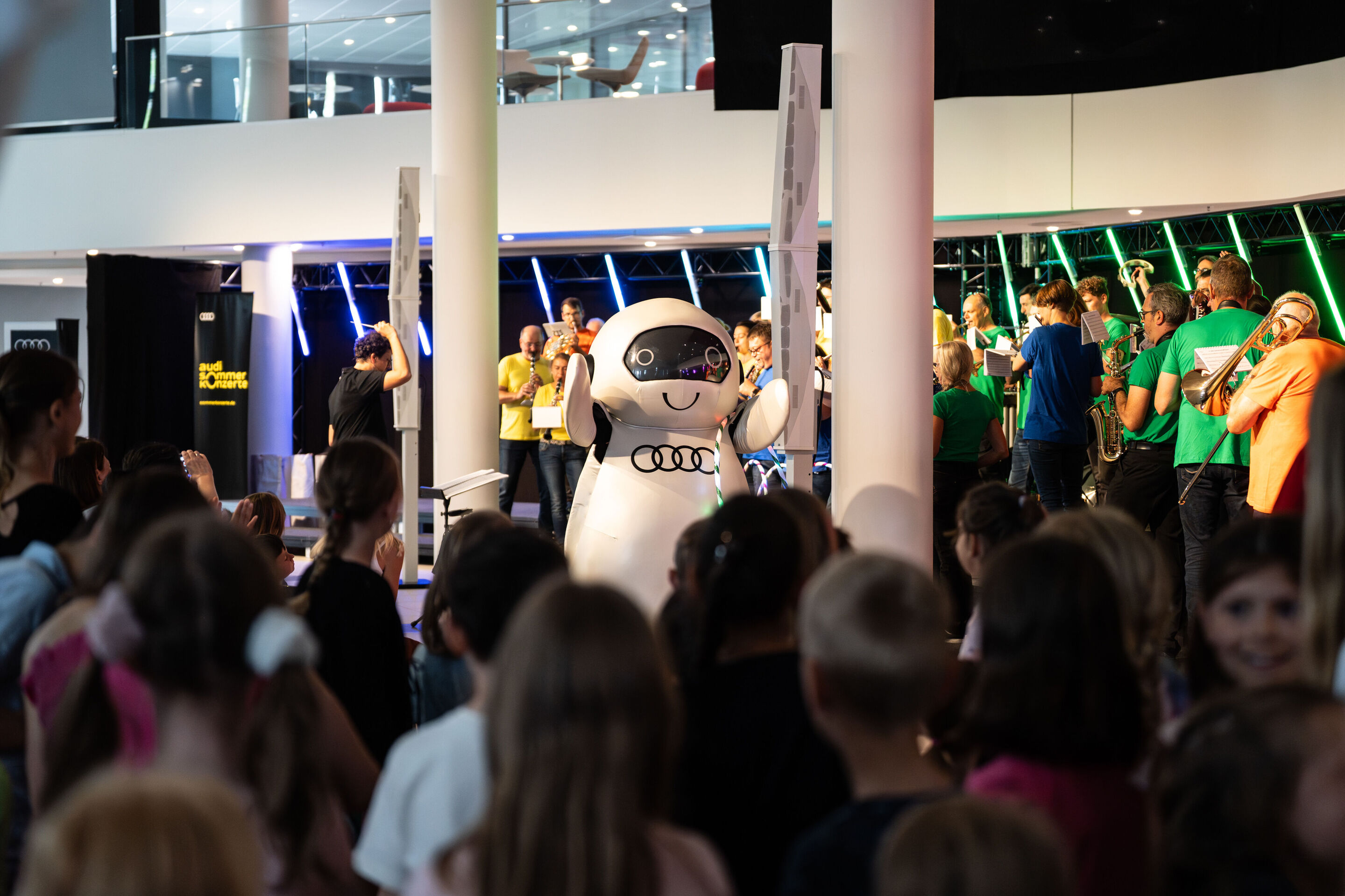 “ADUI und die Kraft der magischen Ringe”: children’s concert with the Audi Philharmonic Wind Orchestra - The robot ADUI, bearing the four Audi rings, stands in front of the stage. In the background, the Audi Philharmonic Wind Orchestra performs under...