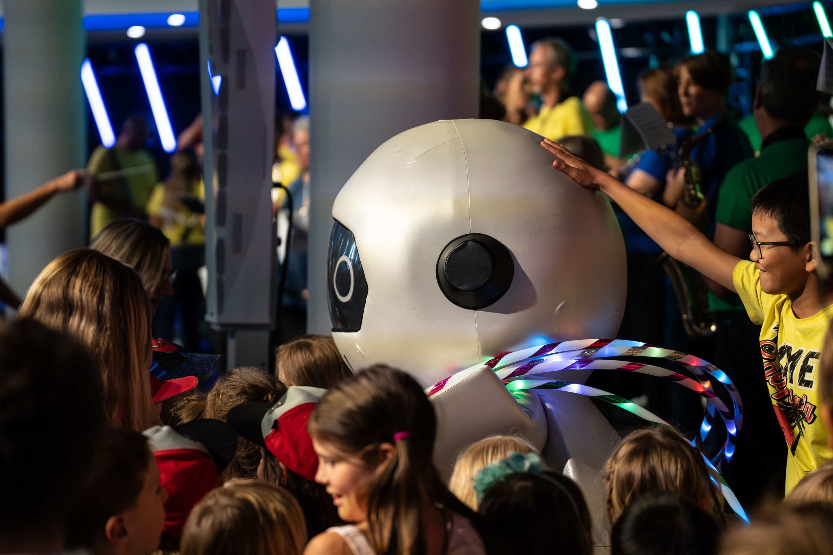 “ADUI und die Kraft der magischen Ringe”: children’s concert with the Audi Philharmonic Wind Orchestra - The robot ADUI, bearing the four Audi rings, stands in front of the stage. In the background, the Audi Philharmonic Wind Orchestra performs under...