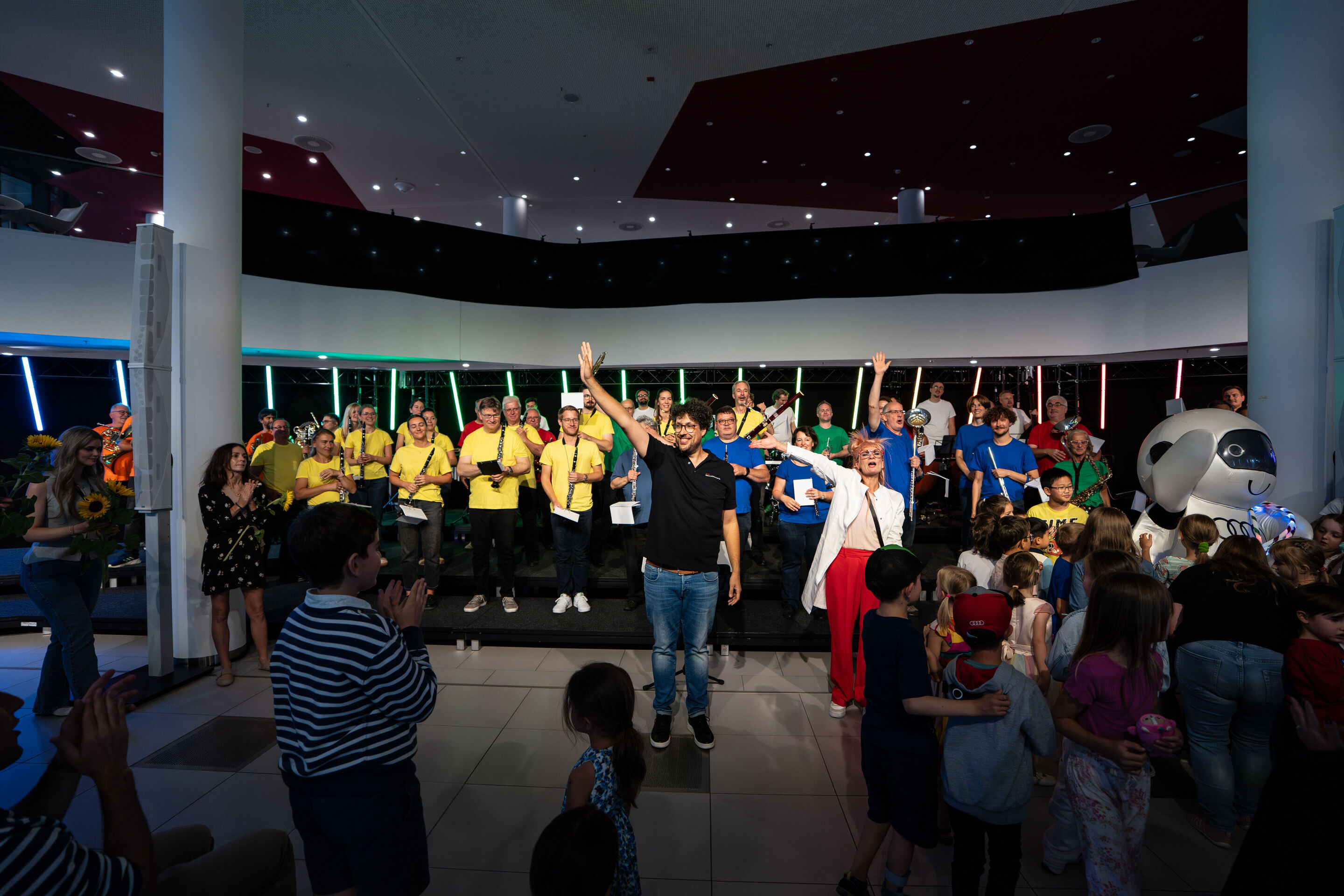 “ADUI und die Kraft der magischen Ringe”: children’s concert with the Audi Philharmonic Wind Orchestra - On stage, the Audi Philharmonic Wind Orchestra stands with conductor Pietro Sarno in the center and actress Olivia Wendt to his right, waving to the...