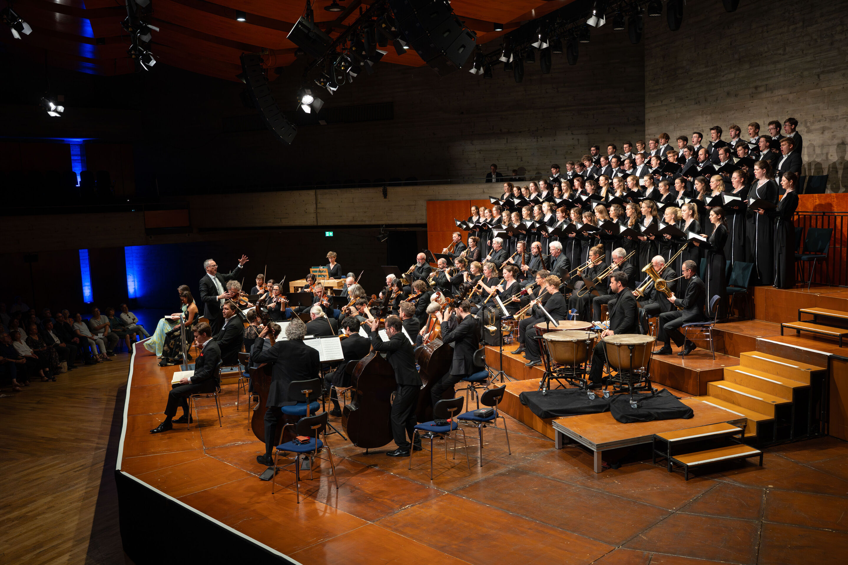 Mendelssohns „Elias“ mit der Audi Jugendchorakademie - Auf der Bühne des Festsaals Ingolstadt dirigiert Martin Steidler links im Bild die Akademie für Alte Musik Berlin. Dahinter steht die Audi Jugendchorakademie mit Notenbüchern in den Händen.