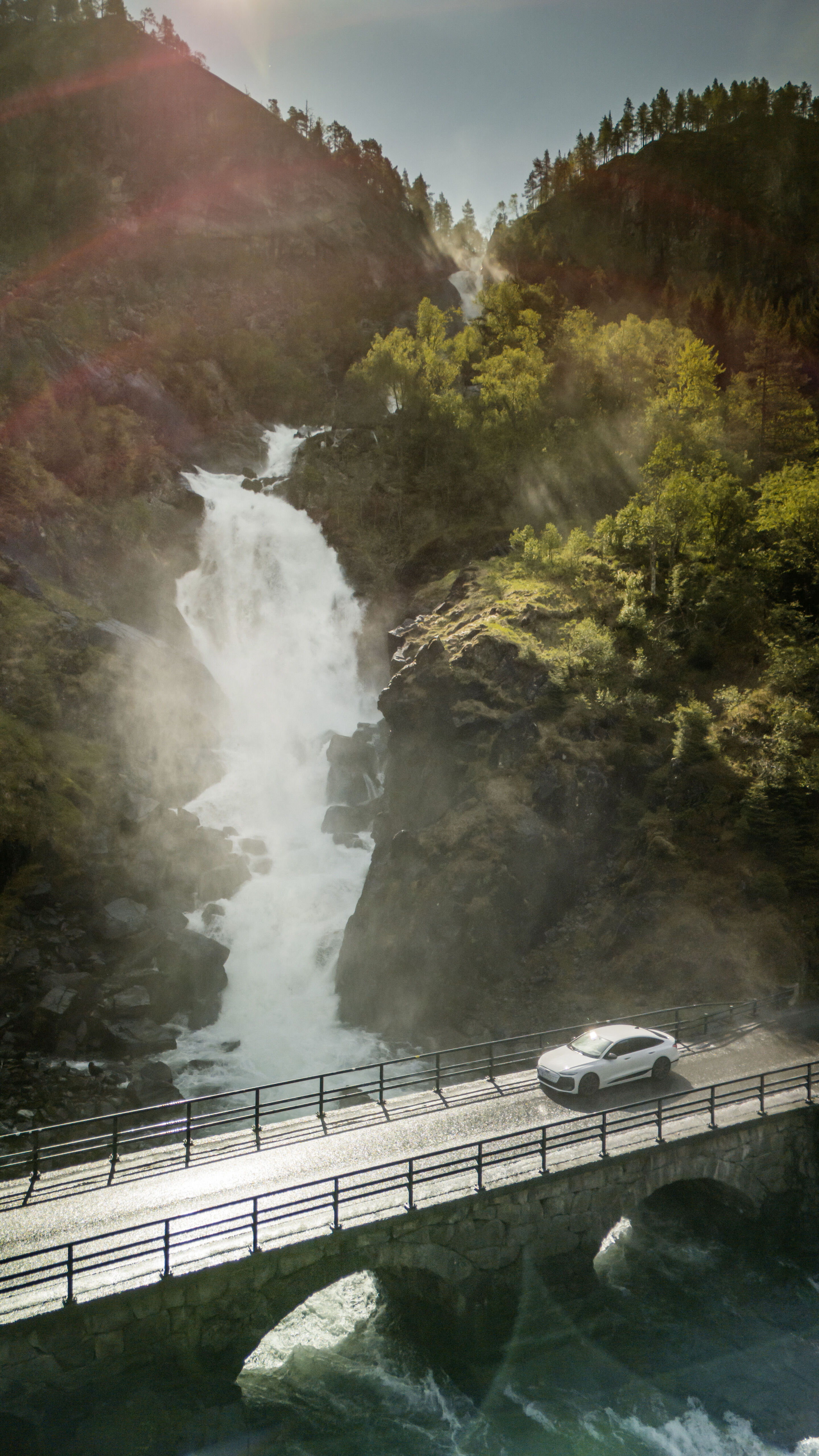 All charged up: from Copenhagen to Bergen in the A6 e-tron - An Audi A6 Sportback e-tron in Glacier white metallic crosses a bridge, with a raging waterfall surrounded by trees in the background.