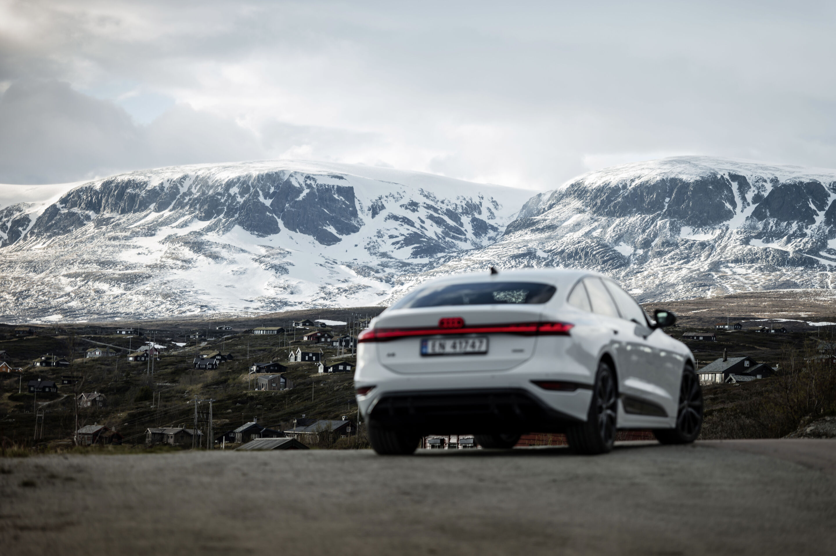 All charged up: from Copenhagen to Bergen in the A6 e-tron - An Audi A6 Sportback e-tron in Glacier white metallic is parked on a road; behind it a Norwegian village with numerous scattered houses and snow-covered mountains are visible.