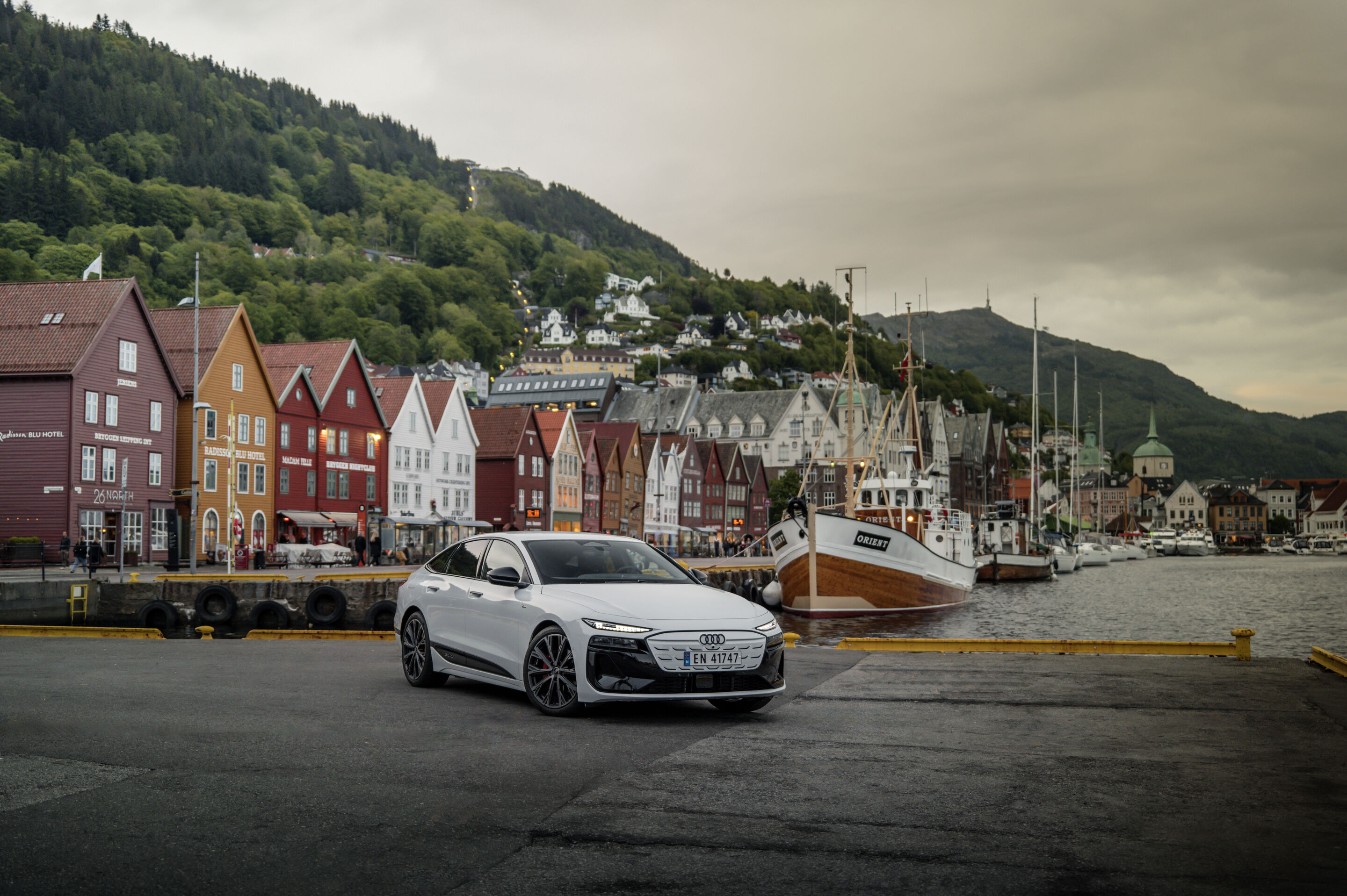 All charged up: from Copenhagen to Bergen in the A6 e-tron - An Audi A6 Sportback e-tron in Glacier white metallic is parked in the harbor of the Norwegian city of Bergen. Historic buildings and a fishing boat are in the immediate background.