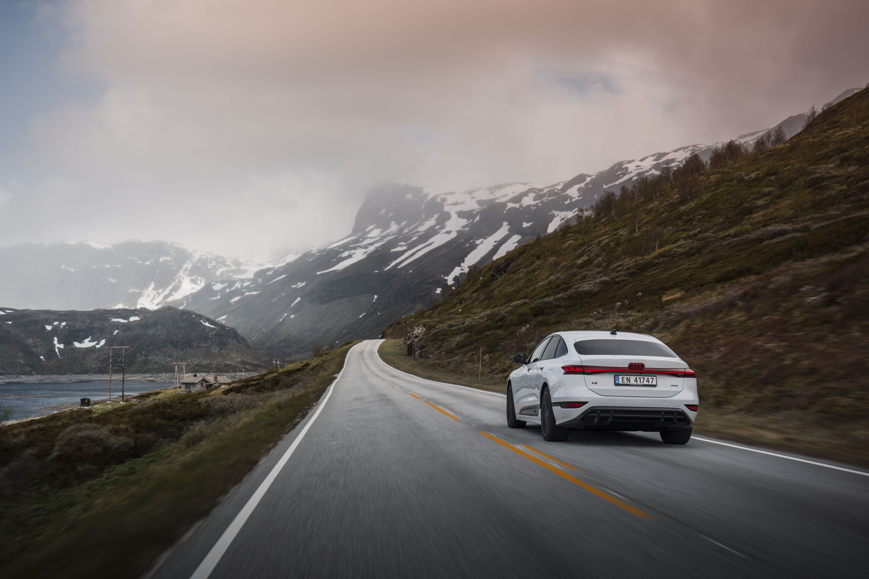 All charged up: from Copenhagen to Bergen in the A6 e-tron - An Audi A6 Sportback e-tron in Glacier white metallic drives along a road in the Norwegian countryside. Snow-covered mountains are in the background.