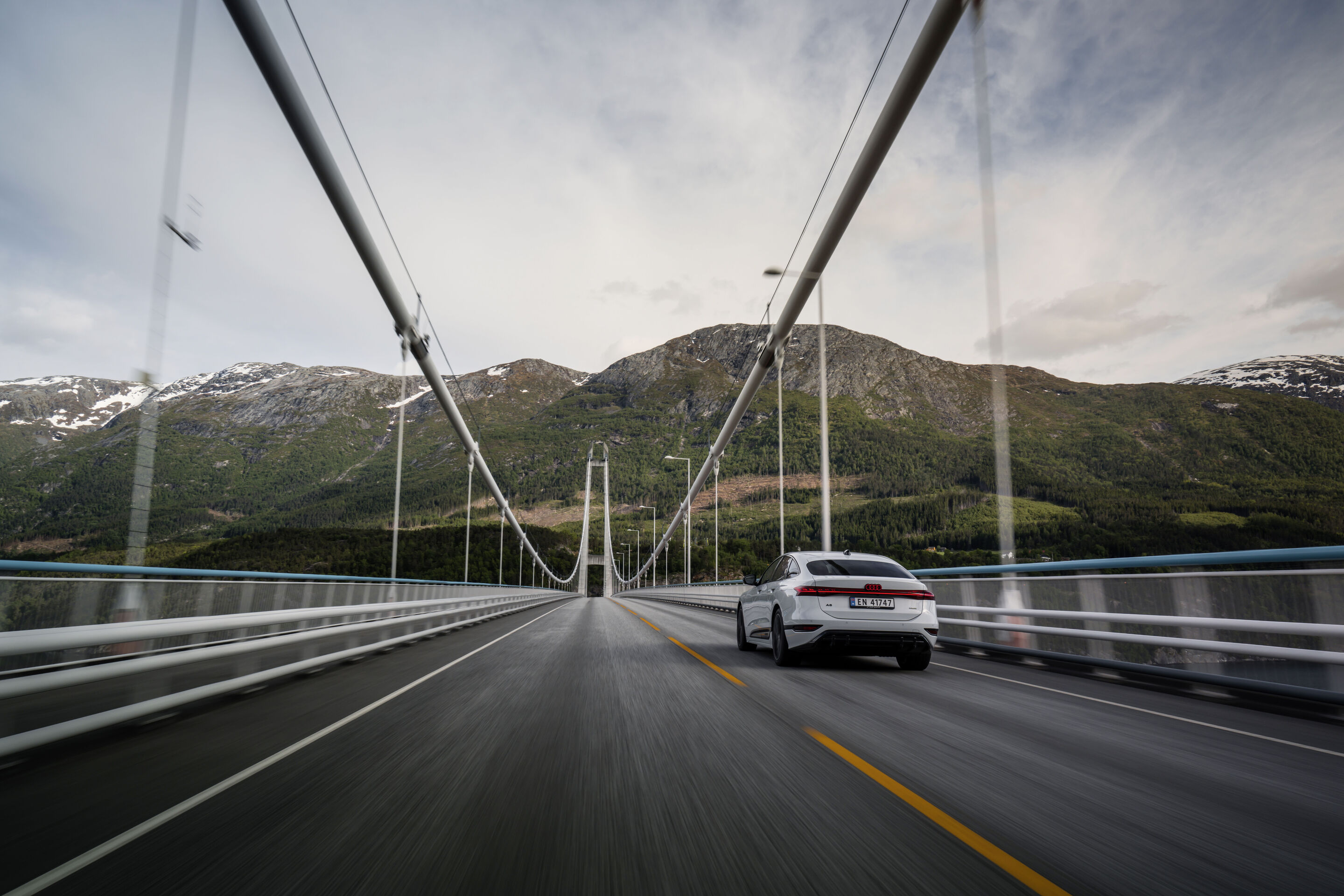 All charged up: from Copenhagen to Bergen in the A6 e-tron - An Audi A6 Sportback e-tron in Glacier white metallic drives over the Hardanger Bridge in Norway. Wooded mountains are visible in the background.