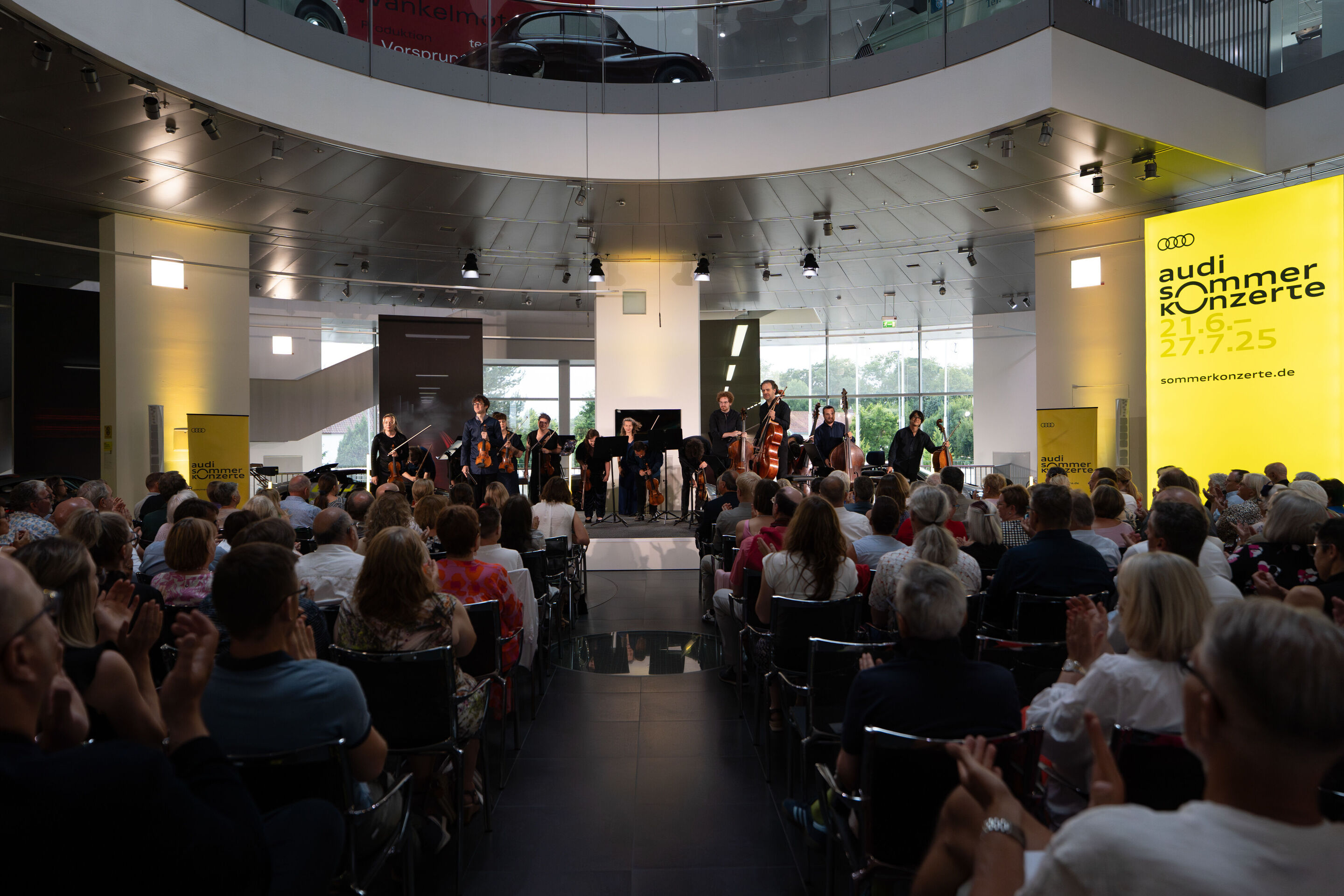 “Crossing over the Atlantic”: from Britten to Nirvana with the O/Modernt chamber orchestra - On the stage of the Audi museum mobile, from the audience's perspective, stands the O Modernt Chamber Orchestra under the direction of Hugo Ticciati.