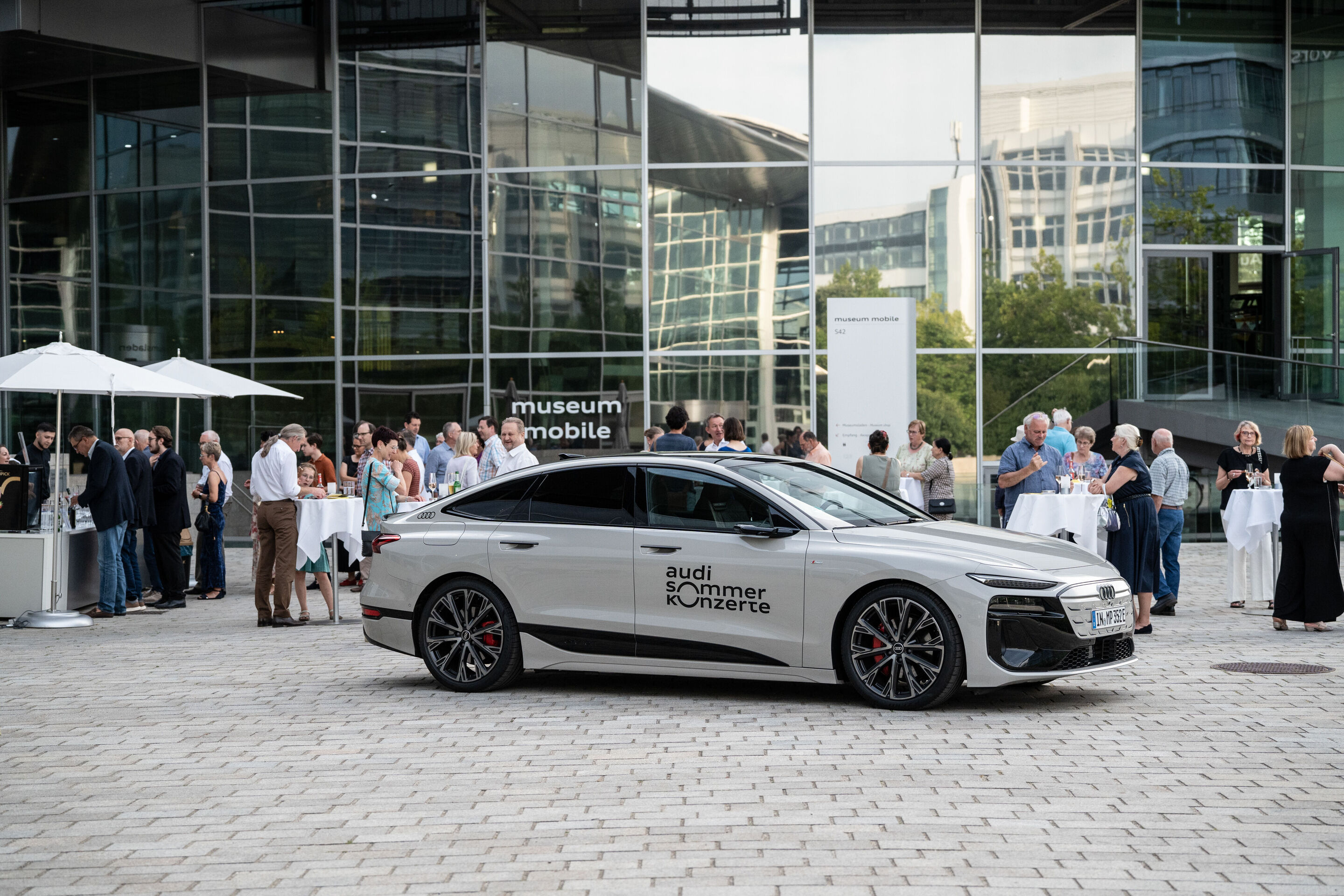 Alinde Quintet – ARD-Musikwettbewerb Preisträger - Ein Audi A6 Sportback e-tron in der Farbe Siambeige Metallic mit dem Logo der Audi Sommerkonzerte auf der Piazza vor dem museum mobile. Im Hintergrund: Gäste der Audi Sommerkonzerte.