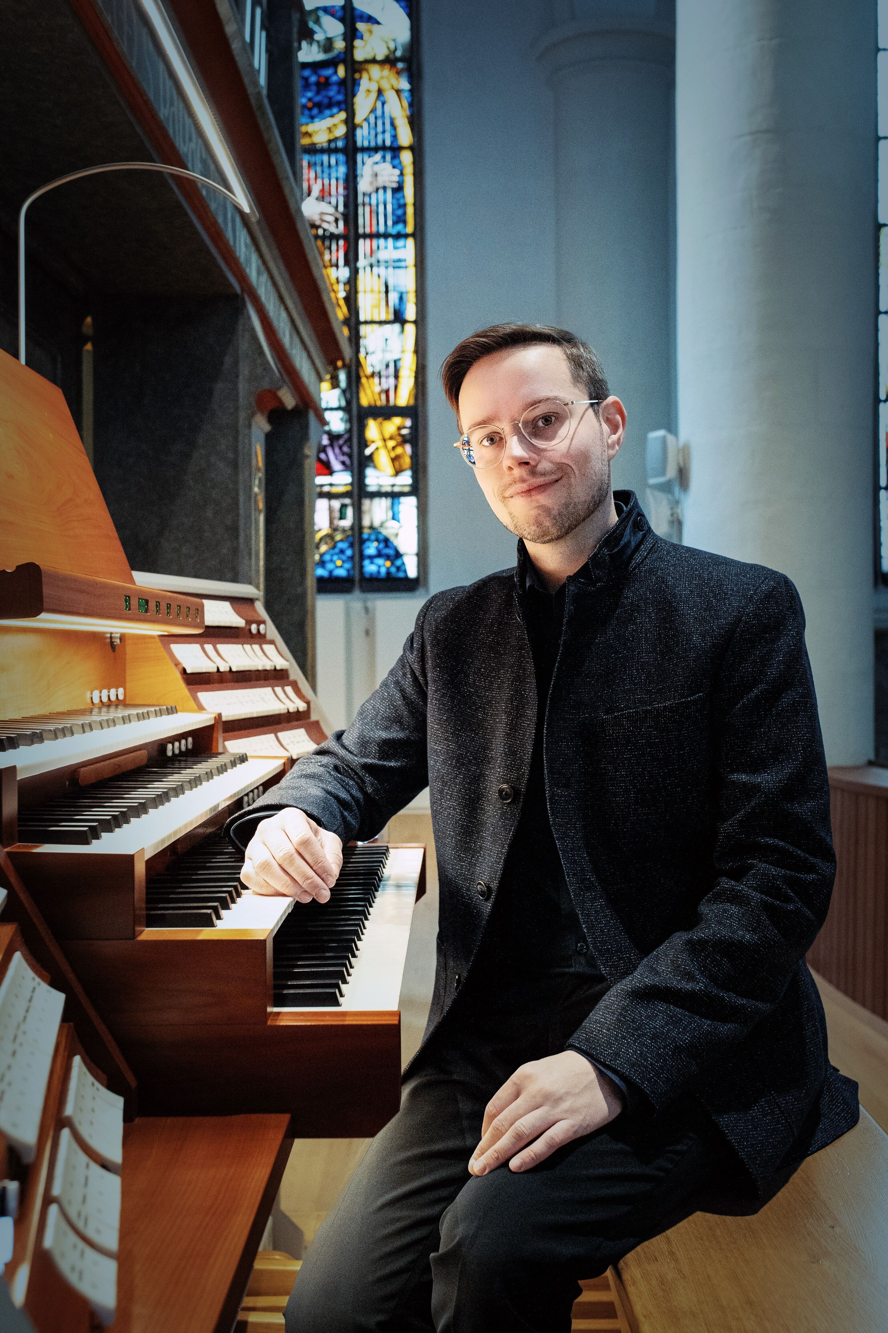 Audi Bläserphilharmonie: Drei Konzert-Highlights im März und Mai - Martin Gregorius sitzt an einer Orgel in einer Kirche. Im Hintergrund sind bunte Kirchenfenster und weiße Säulen zu sehen.