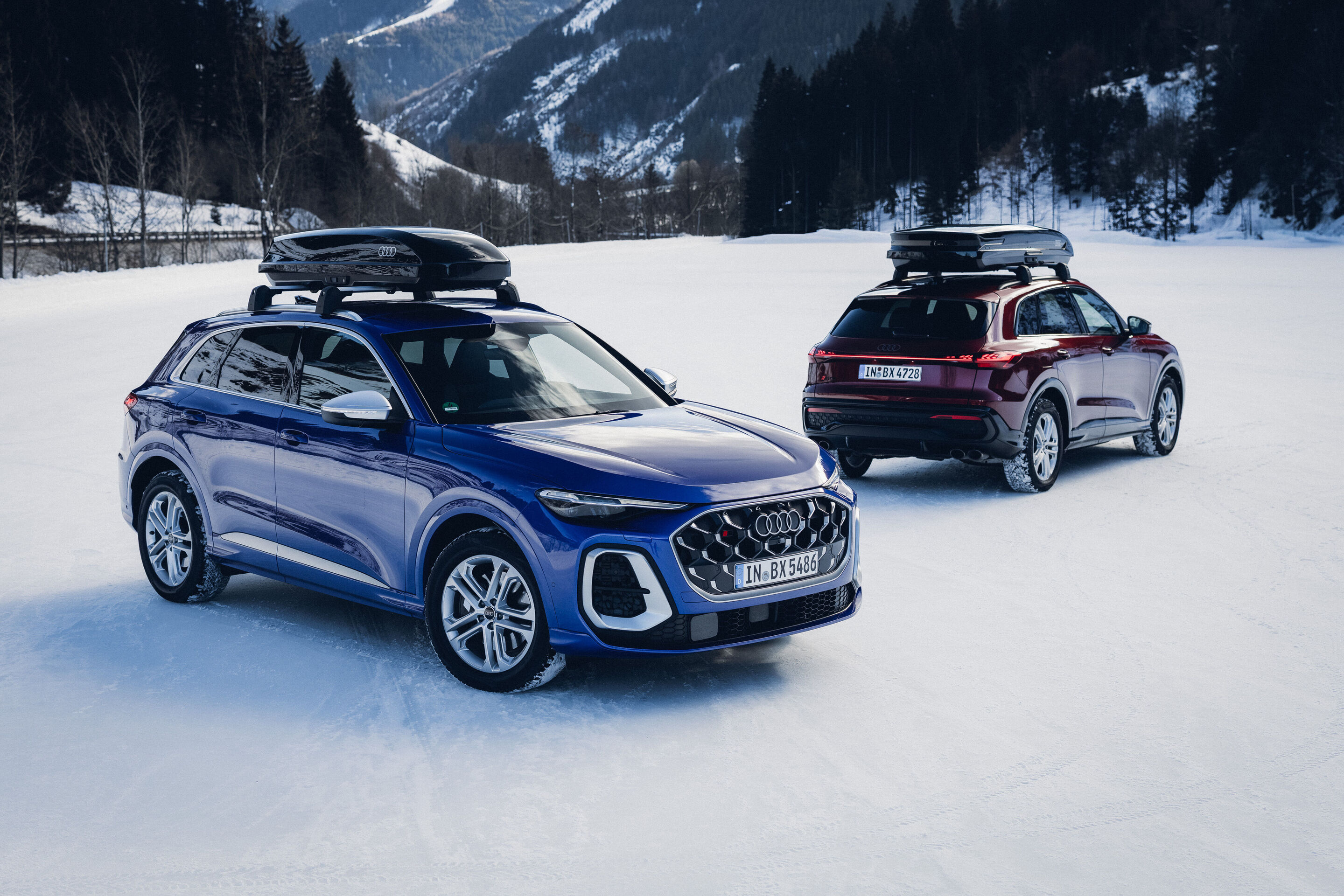 Audi SQ5 - Two Audi SQ5 are parked on a snowy open area in the mountains. Both vehicles have roof cargo boxes; one is Ultra Blue and shown from the front, while the other is Grenadine Red and viewed from the rear.