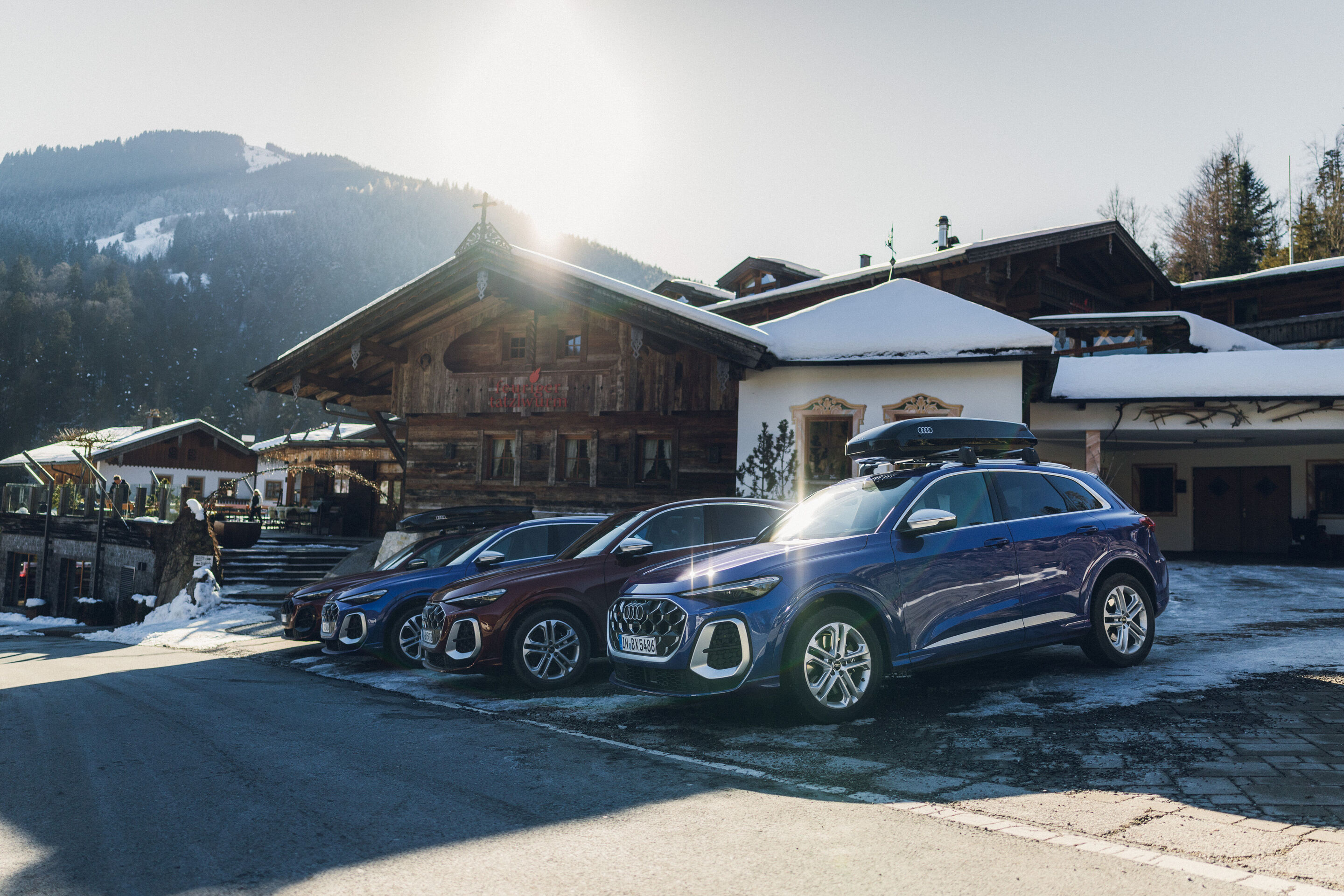 Audi SQ5 and Audi SQ5 Sportback - Four Audi SQ5 models are parked side by side in front of a traditional alpine wooden building. The vehicles—in Ultra blue, Grenadine red, and Daytona grey—are on a partially snowy driveway, the frontmost and the rearmost...