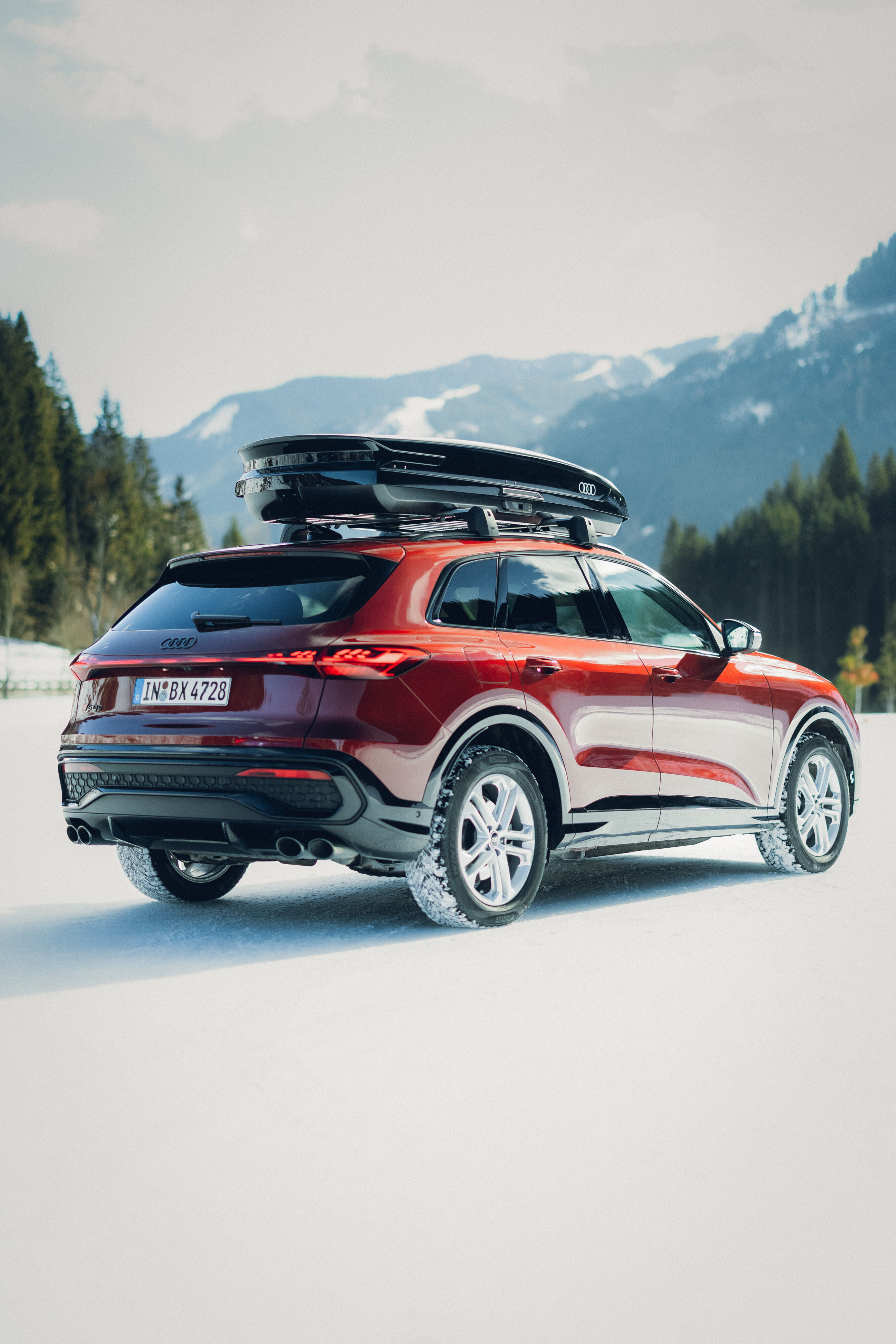 Audi SQ5 - An Audi SQ5 in Grenadine Red is parked on a snowy surface with mountains in the background. The vehicle is shown from the rear and side and is equipped with a black roof cargo box.