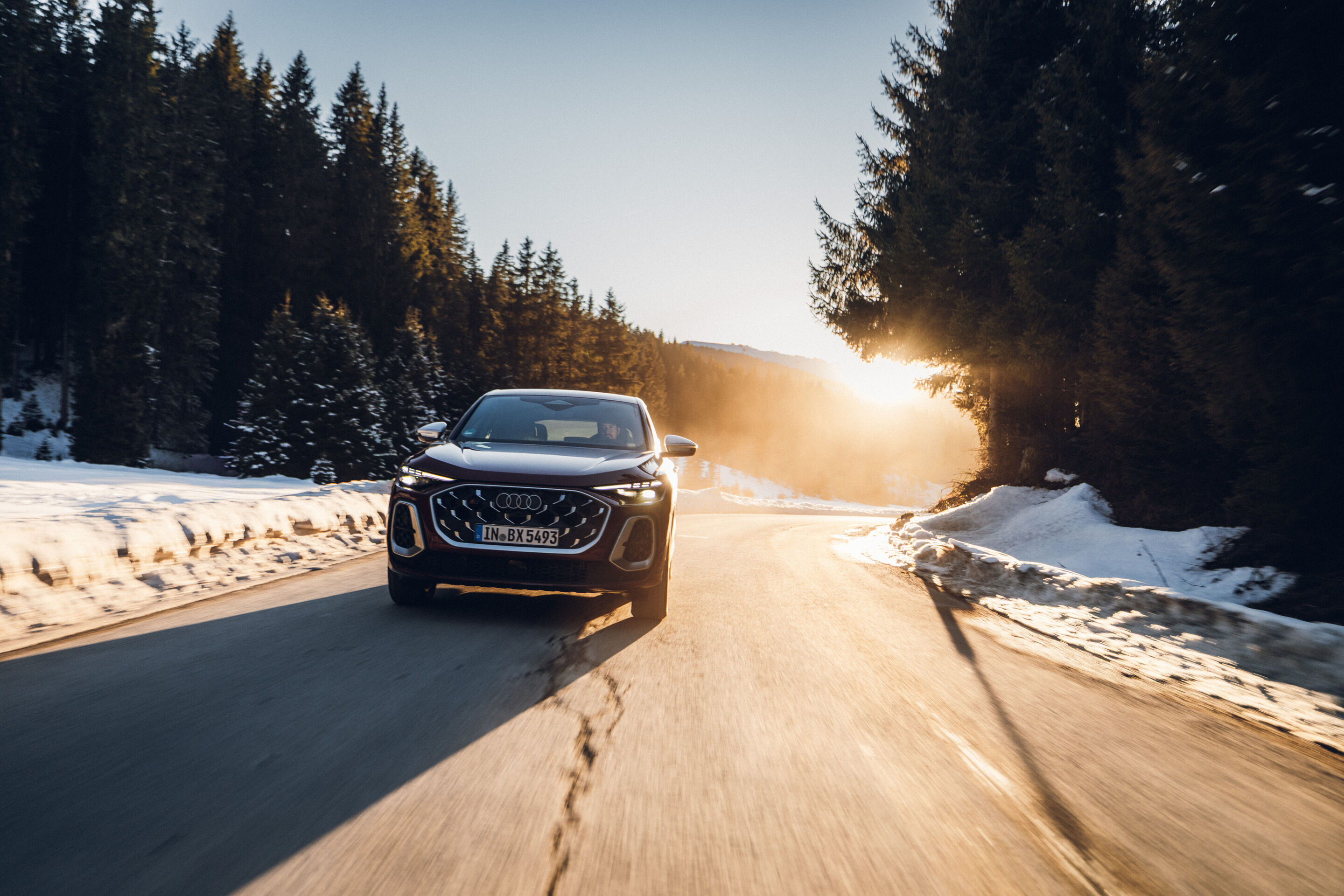 Audi SQ5 Sportback - An Audi SQ5 Sportback in Grenadine Red is driving along a winter mountain road. Snow lines the roadside, and the low sun shines through the trees.