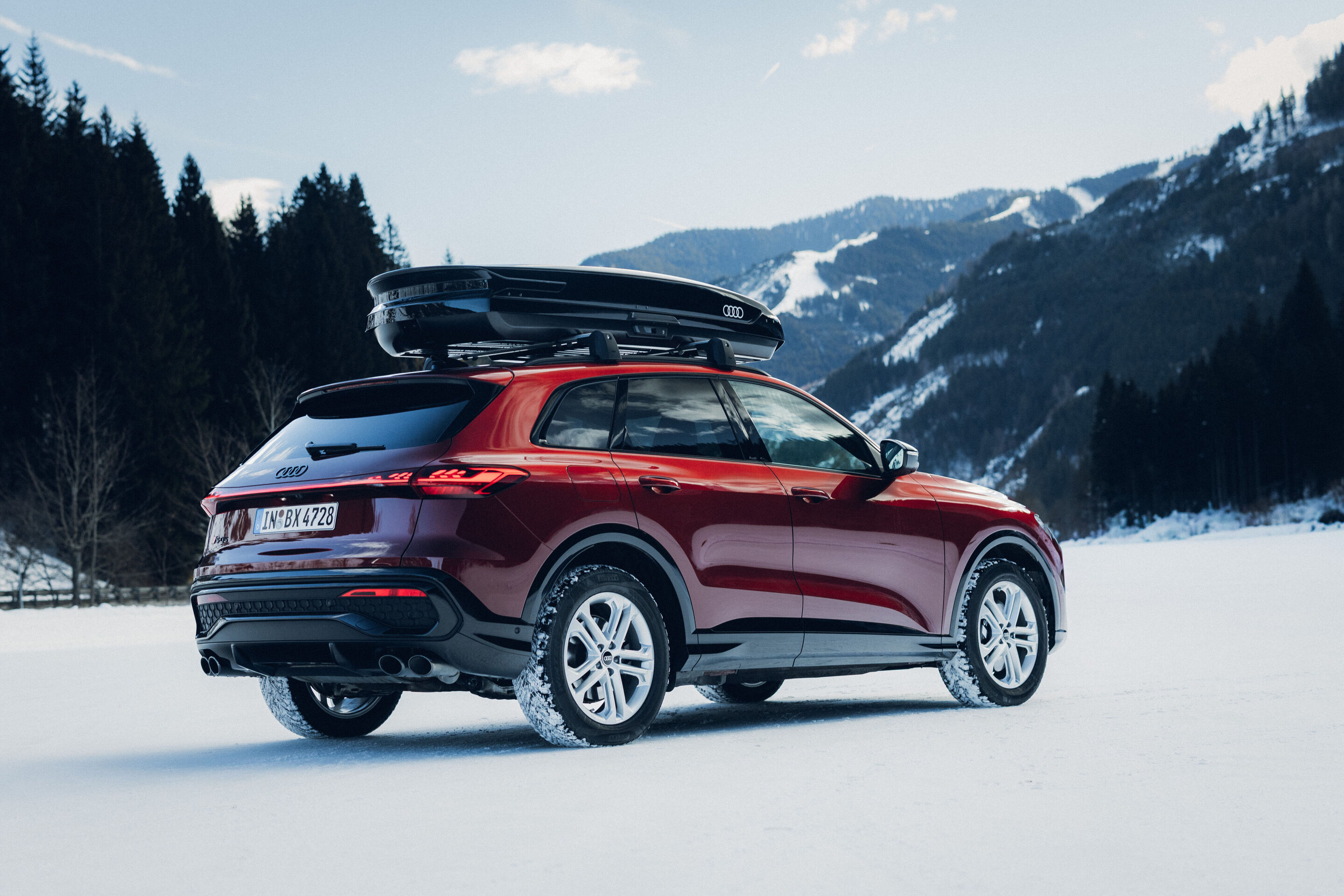 Audi SQ5 - An Audi SQ5 in Grenadine Red is parked on a snowy surface with mountains in the background. The vehicle is shown from the rear and side and is equipped with a black roof cargo box.