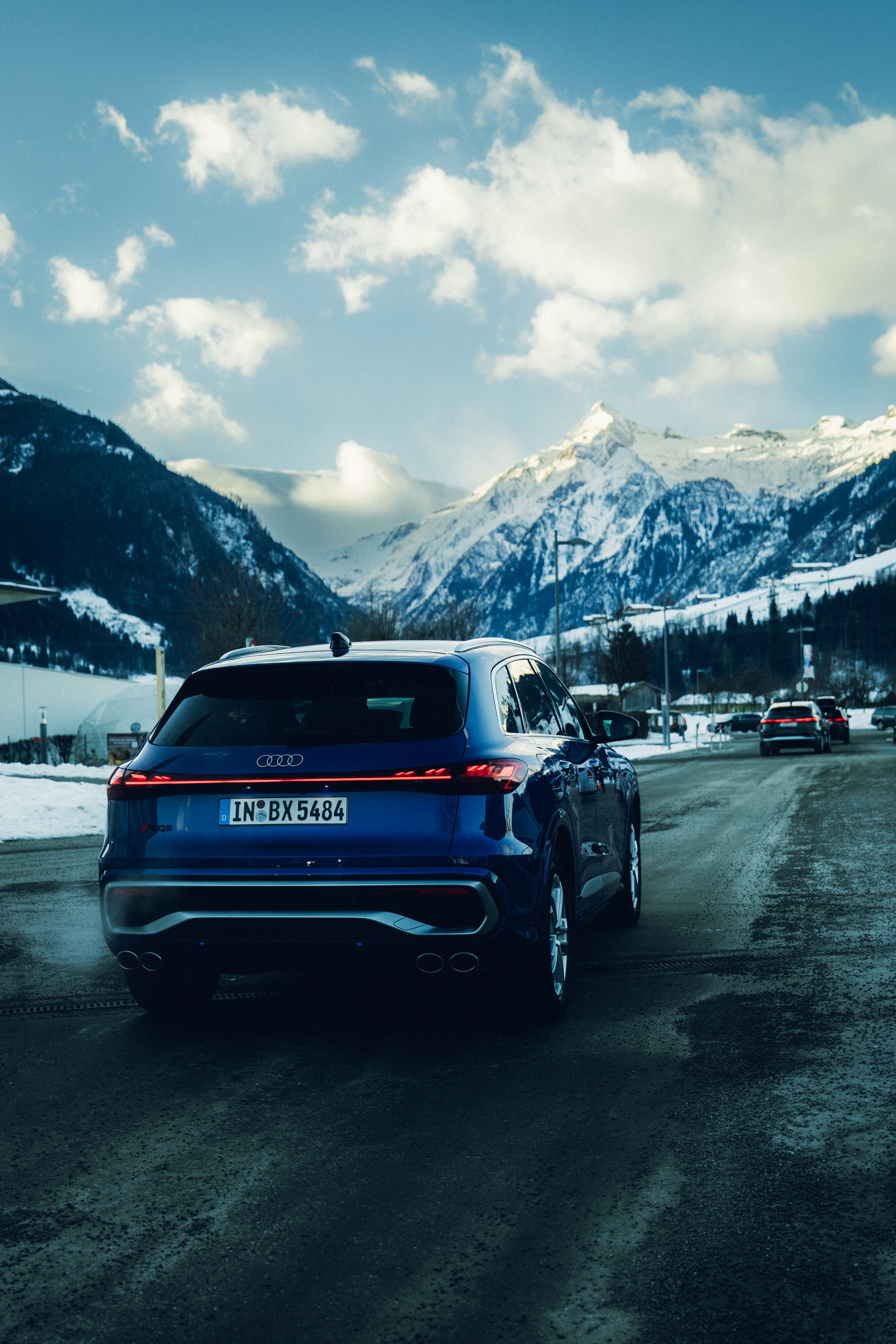 Audi SQ5 - An Audi SQ5 in Ultra Blue is driving along a winter country road, seen from the rear. Snow-covered mountains with high peaks and a partly cloudy sky rise in the background. Two additional vehicles are visible farther ahead on the road.