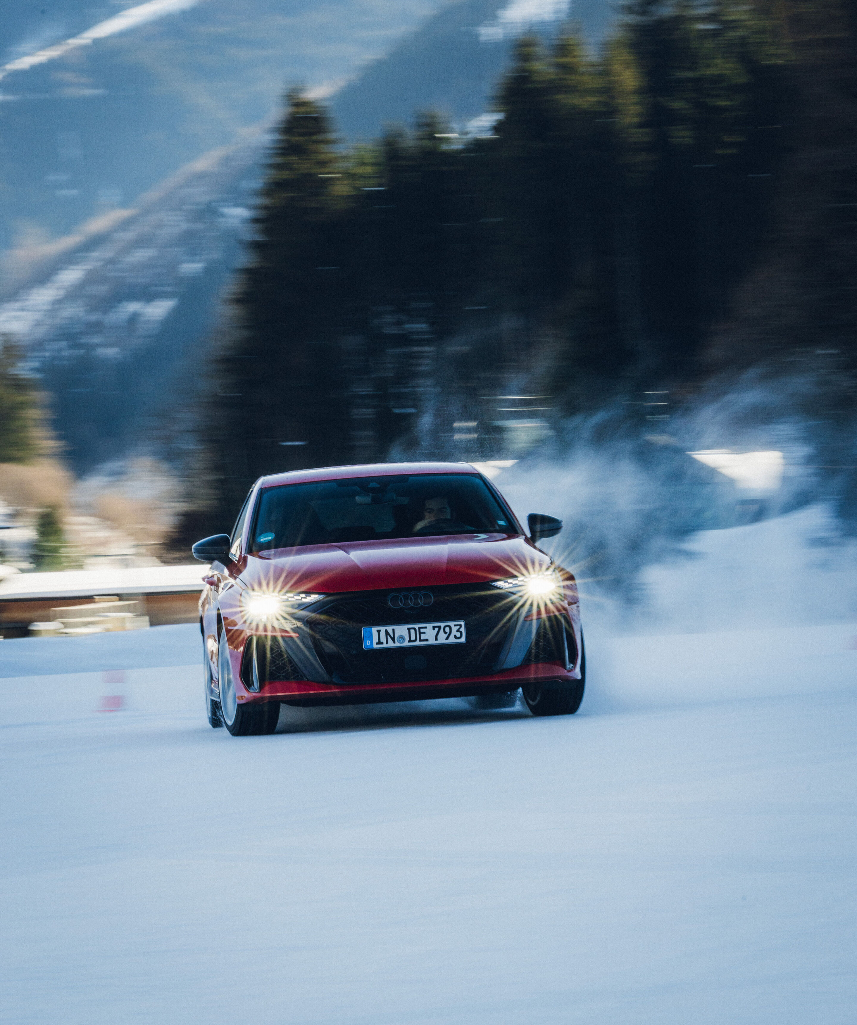 Audi RS 3 Sportback - An Audi RS 3 Sportback in Progressive Red is driving across a snowy surface, kicking up snow. The car is shown from the front with its headlights on.