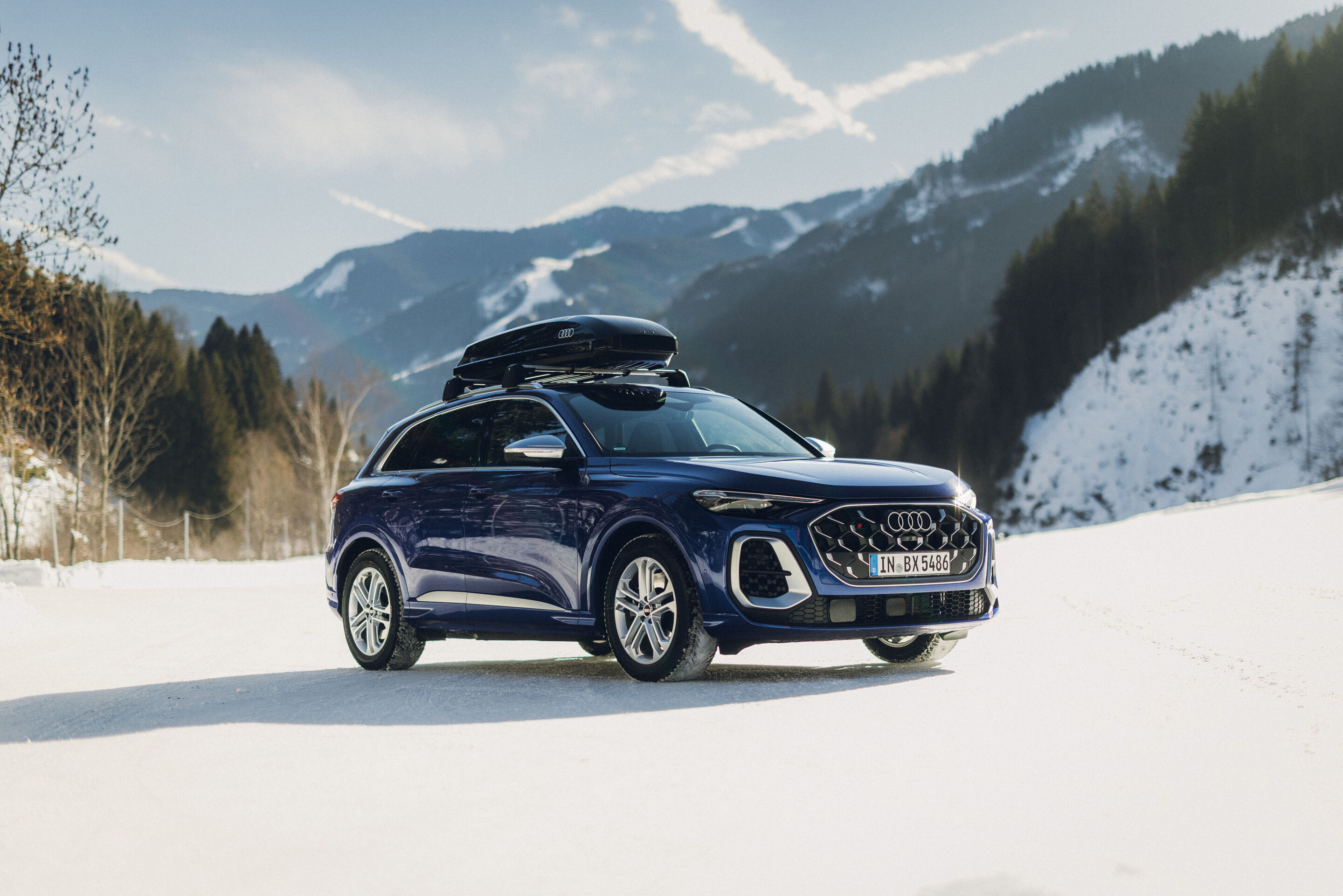 Audi SQ5 - An Audi SQ5 in Ultra Blue is parked on a snowy surface with mountains in the background. The vehicle is shown from the rear and side and is equipped with a black roof cargo box.
