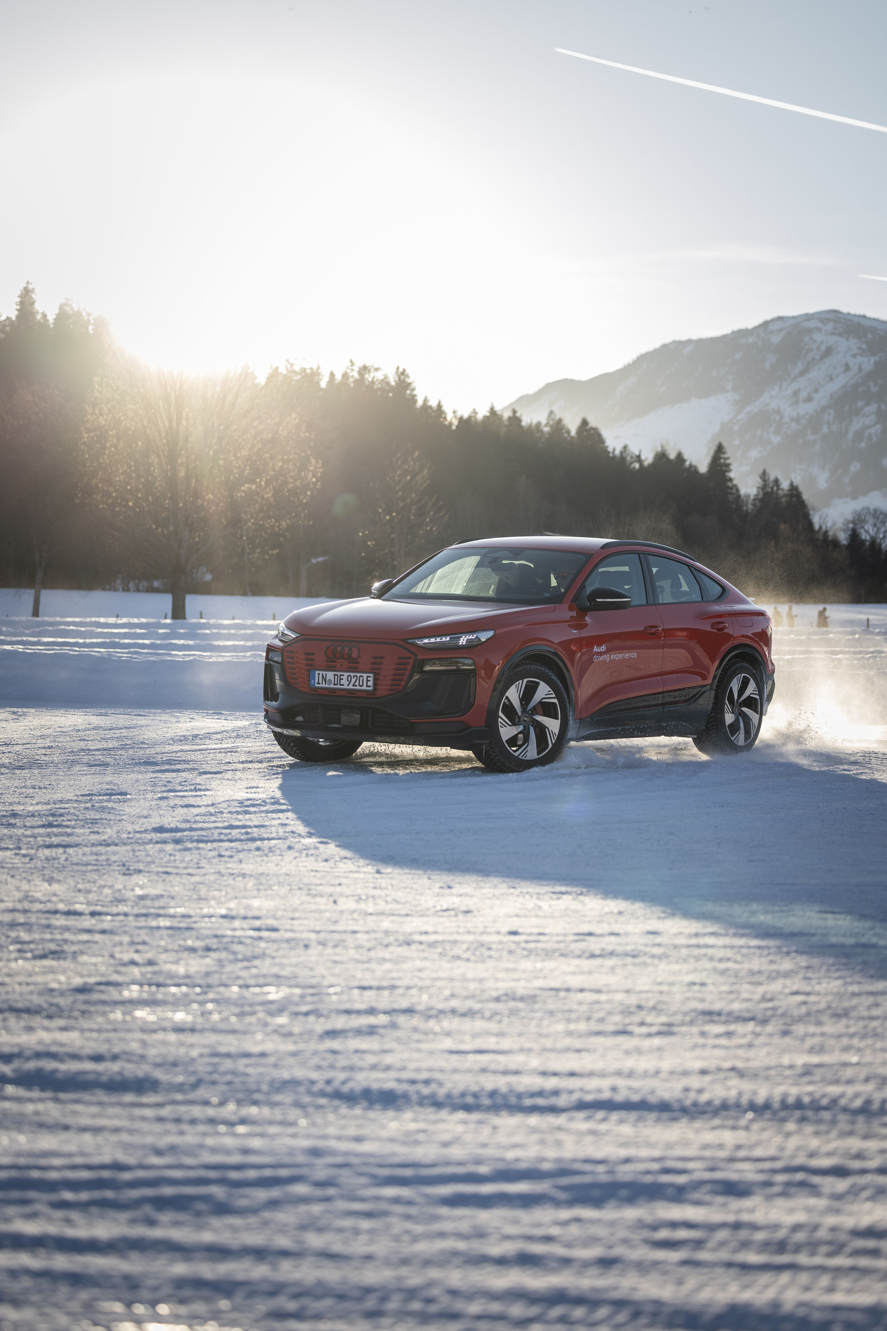 Audi Q6 Sportback e-tron - The Audi Q6 Sportback e-tron drives across a snow-covered surface, with a forest and snow-covered mountains in the background.
Color: Progressive Red Metallic, outdoor driving shot, exterior, three-quarter front view.