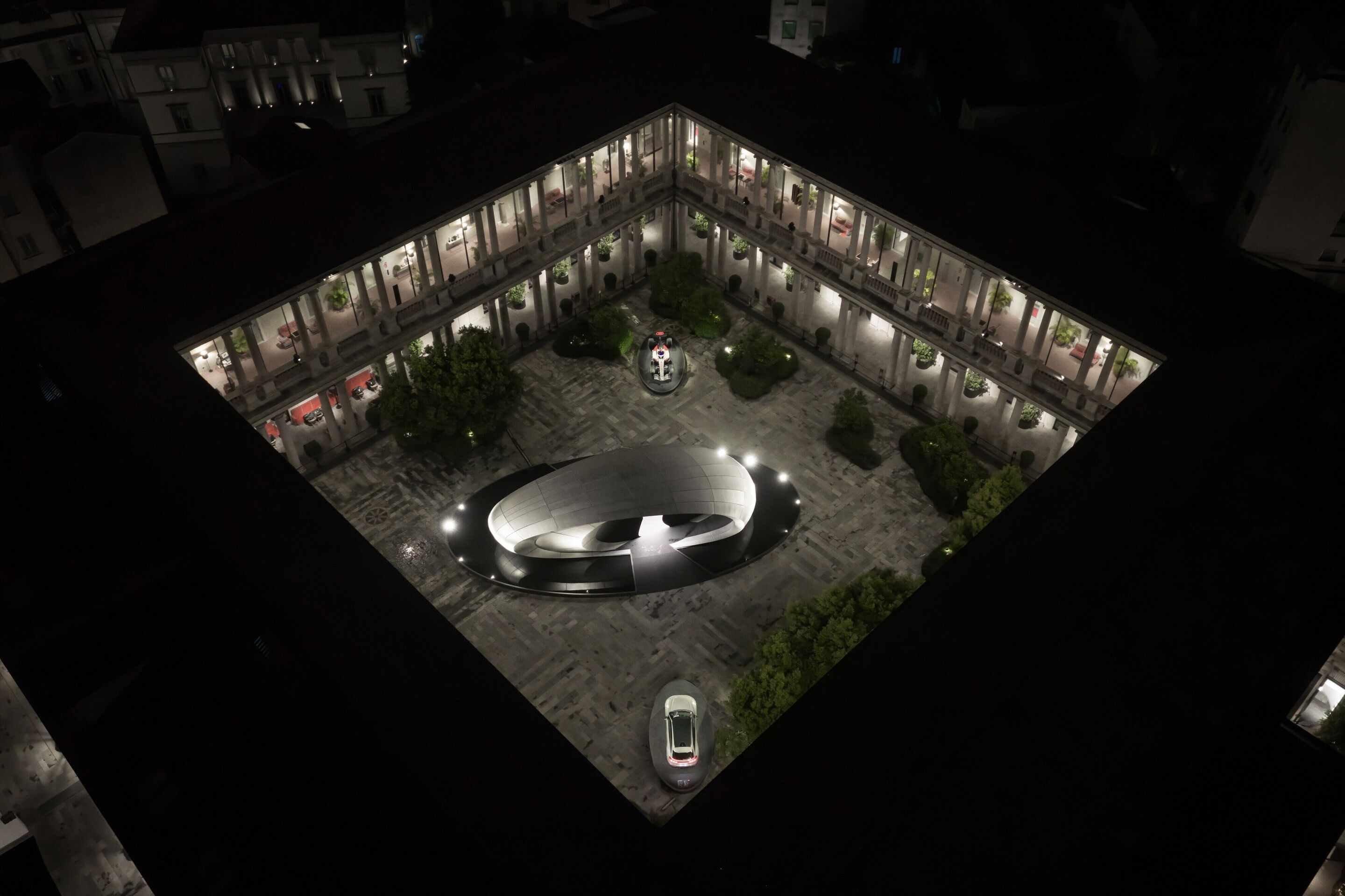 Audi at Milan Design Week 2026 - Night-time aerial view of the installation “Origin” during Milan Design Week 2026. The reduced, oval architectural form contrasts with the historic courtyard architecture, underscoring the approach to design as a cultural...