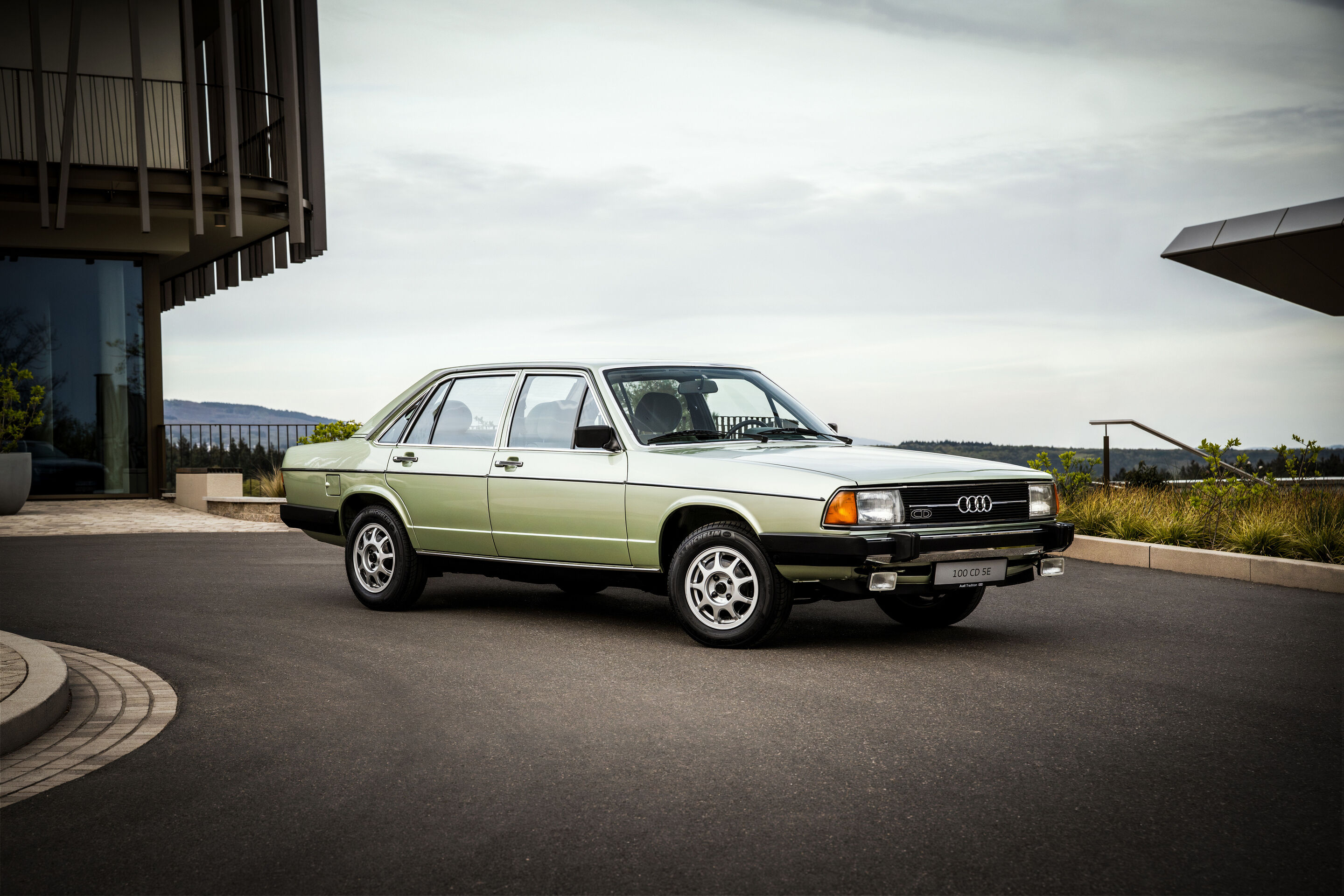50 years of the Audi five-cylinder - An Audi 100 CD (C2, 1979) finished in light metallic green is parked on a paved forecourt in front of modern architecture, shown in side view.