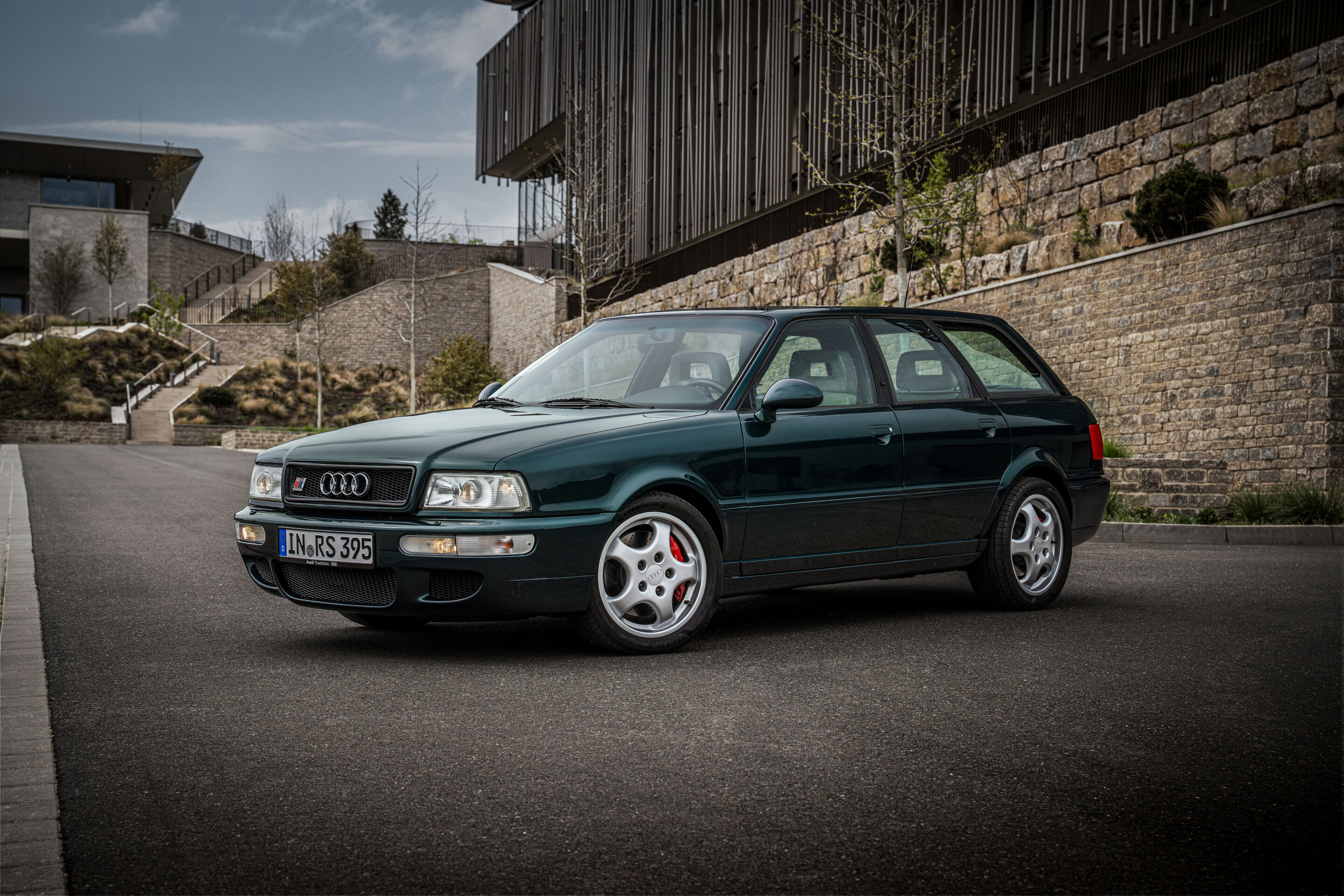 50 years of the Audi five-cylinder - A dark green Audi RS2 Avant from 1995 is positioned on a curved driveway, shown in a three-quarter front view against a stone wall.