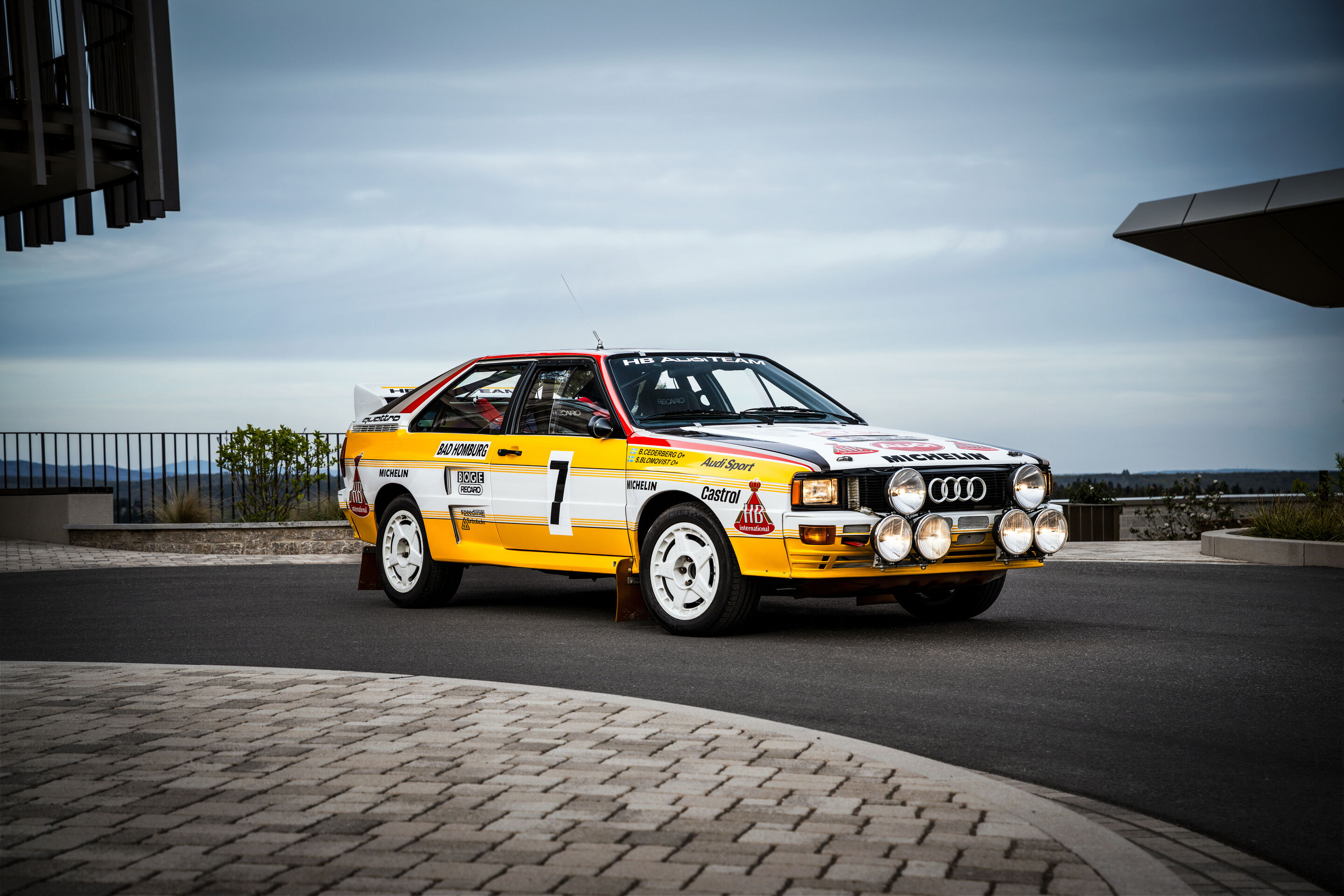 50 years of the Audi five-cylinder - A 1984 Audi rally quattro A2 in yellow-and-white livery with auxiliary headlights is parked on a forecourt.