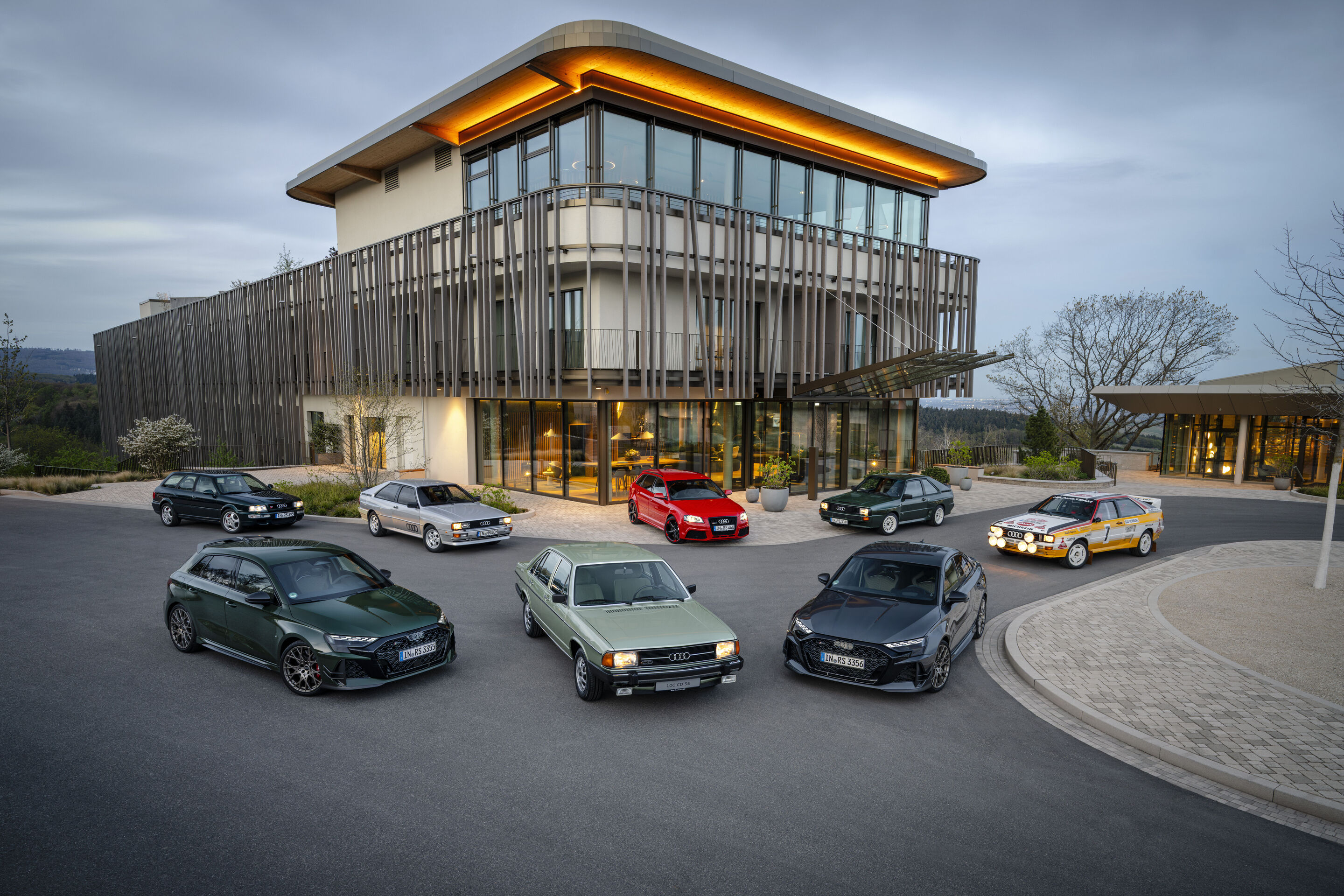 50 years of the Audi five-cylinder - Several Audi RS models from different generations with five-cylinder engine are arranged in a semicircle in front of a modern glass building.
