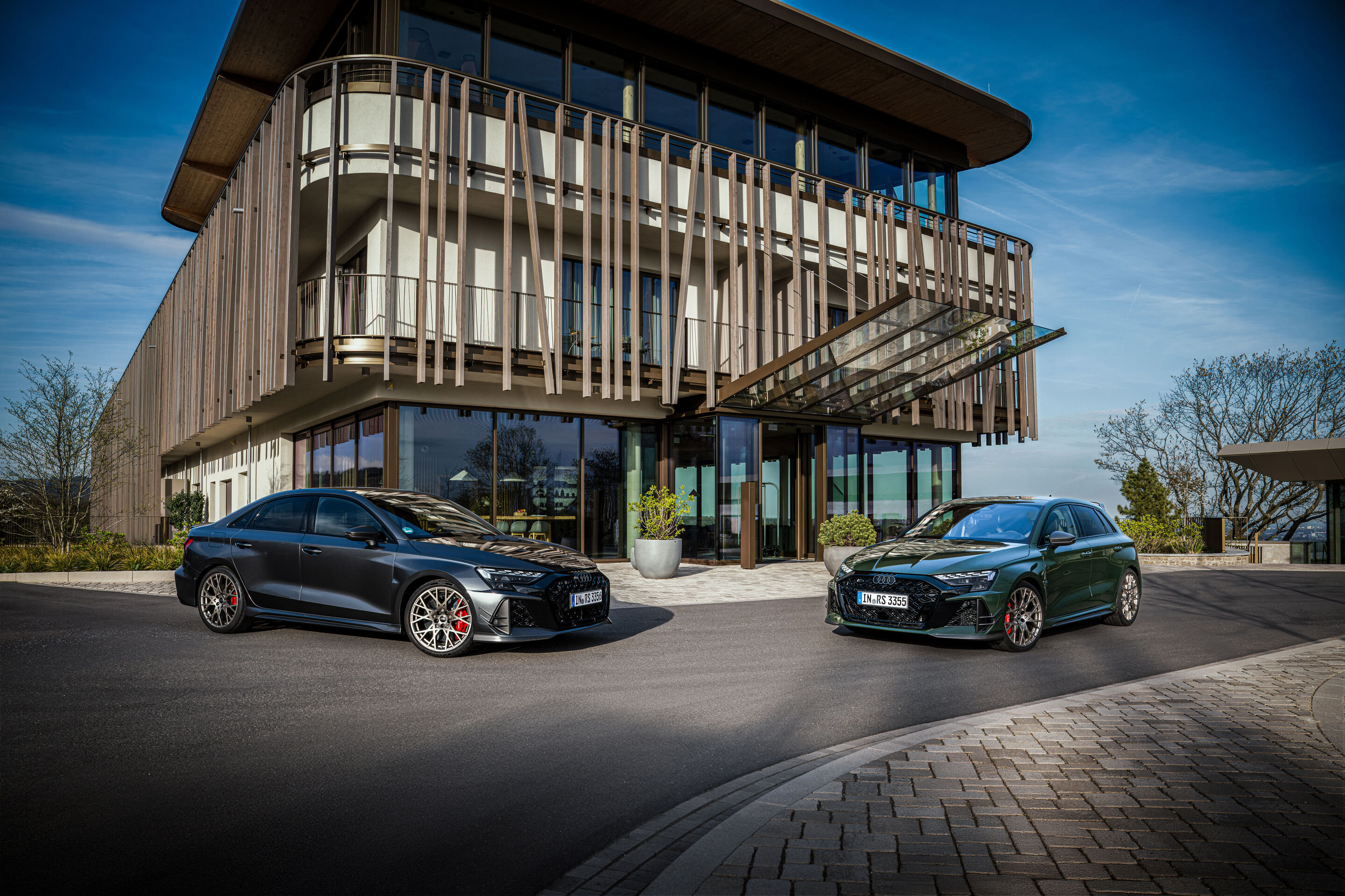 50 years of the Audi five-cylinder - Two Audi RS3 competition models in Malachite Green and Daytona Grey pearl effect are parked facing forward in front of a contemporary building.