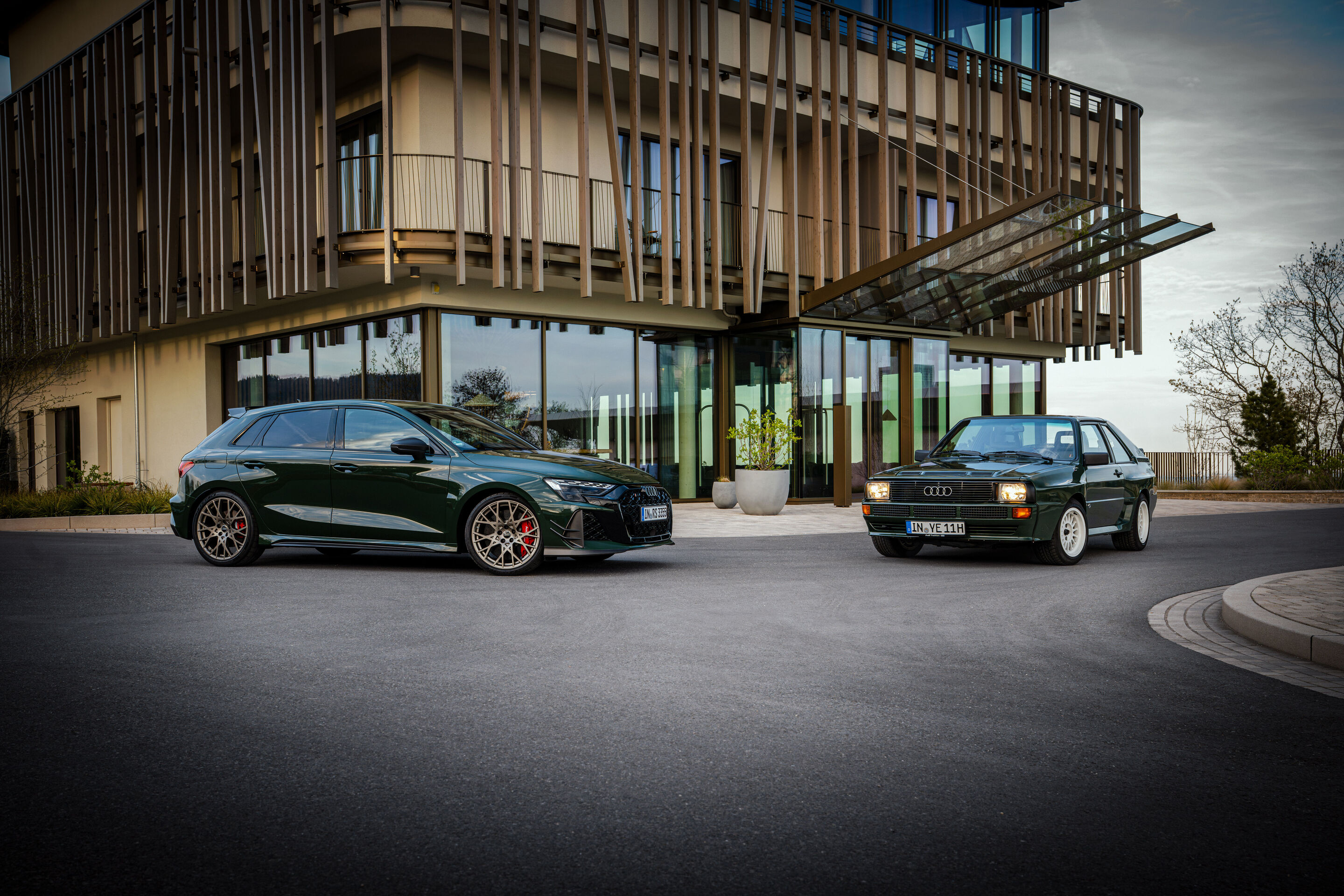 50 years of the Audi five-cylinder - An Audi RS3 competition limited is parked next to a historic 1984 Audi Sport quattro in front of contemporary architecture.