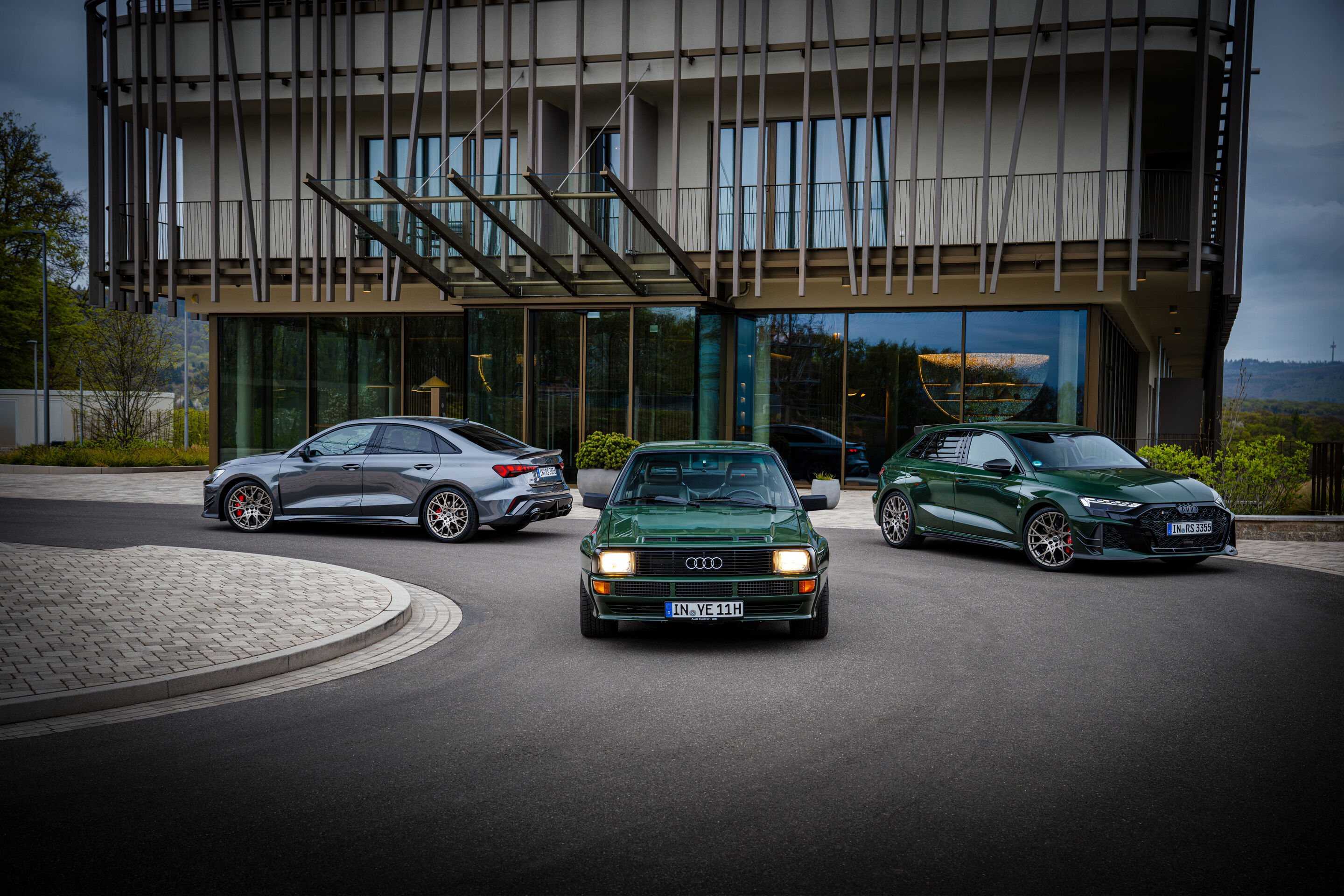 50 years of the Audi five-cylinder - The Audi RS 3 competition limited as a Sedan and Sportback alongside a historic 1984 Audi Sport quattro in front of contemporary architecture.