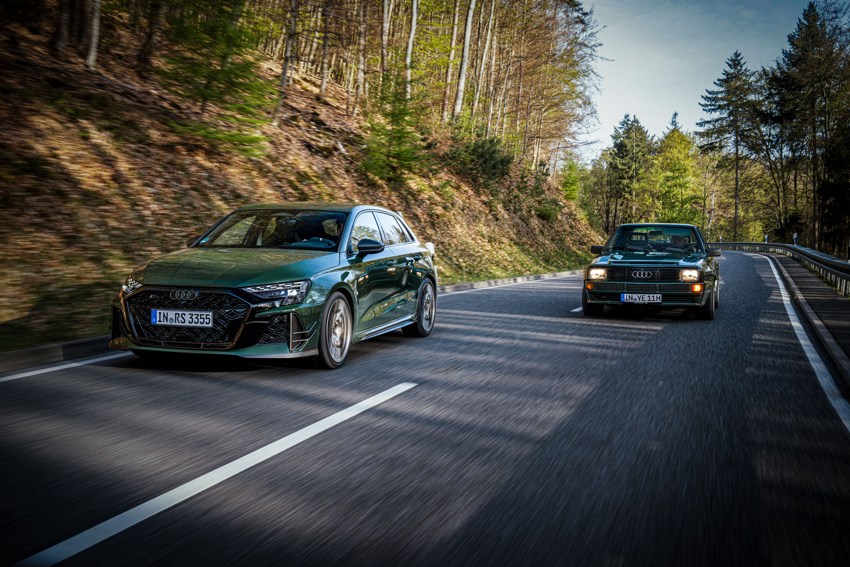 50 years of the Audi five-cylinder - An Audi RS3 competition limited drives ahead of a historic Audi Sport quattro on a winding road through forested landscape.