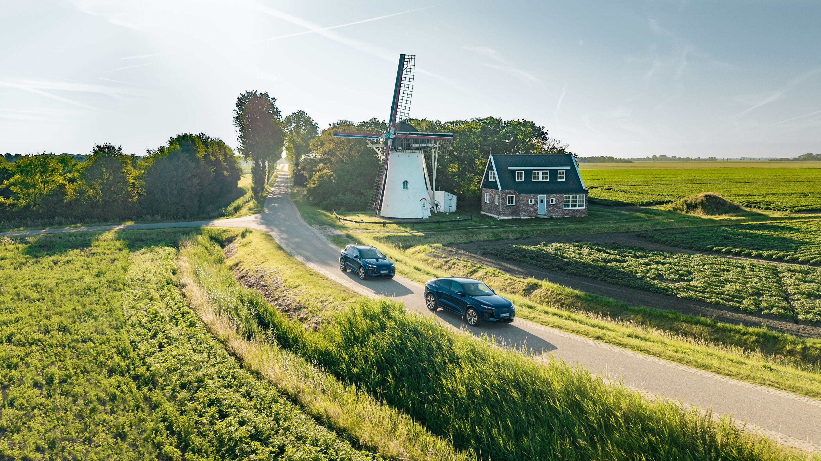 Audi Q6 e-tron Lifecycle Experience - Zwei Audi Q6 e-tron (Ascariblau) fahren hintereinander auf einer schmalen Landstraße durch eine grüne Kulturlandschaft mit Windmühle und Wohnhaus im Hintergrund.